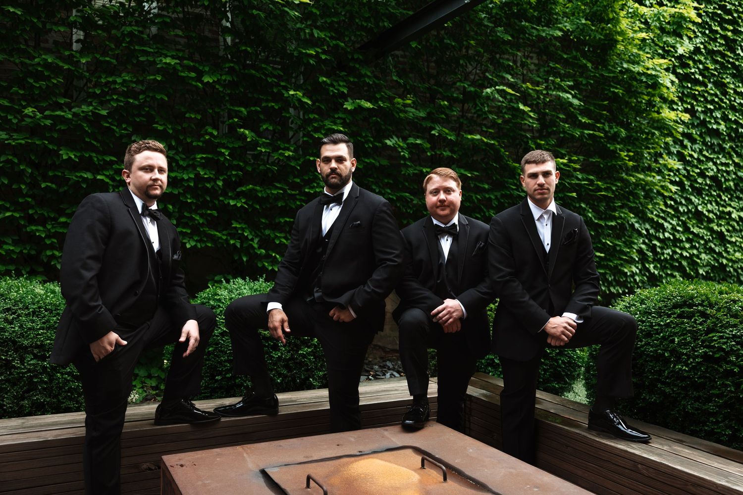 Four groomsmen in black tuxedos posing casually near a fire pit with green ivy backdrop.