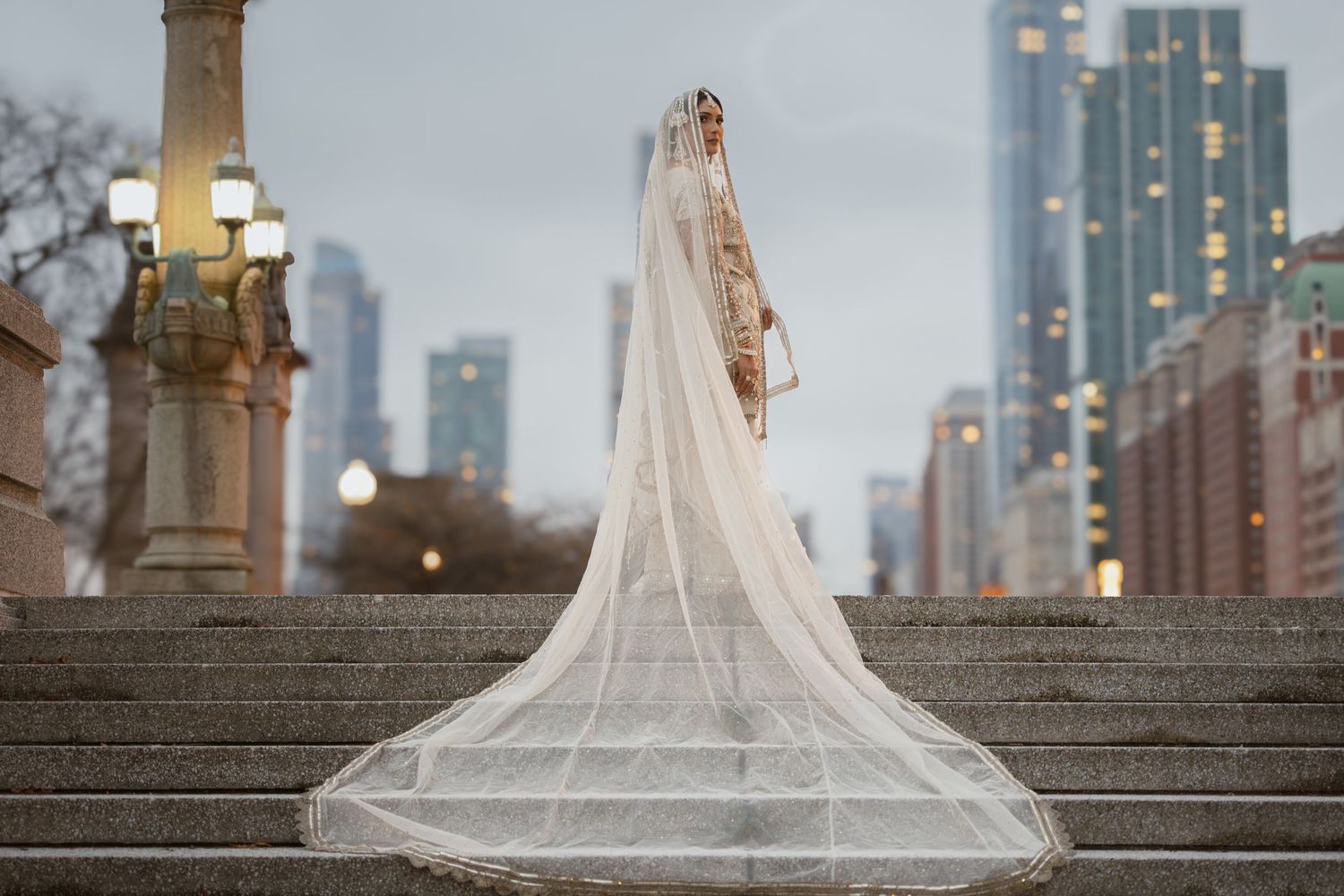 Indian bride in ornate ivory bridal outfit with flowing cathedral veil on stone steps with Chicago skyline.