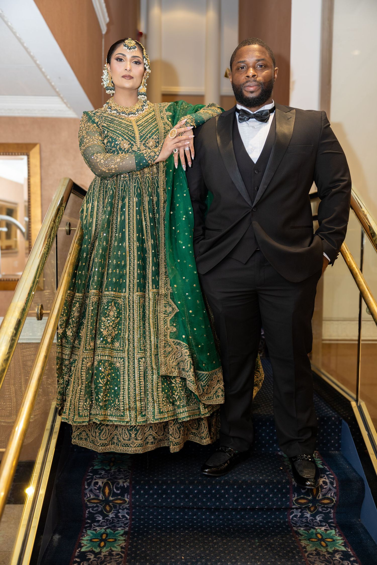 Bride in ornate green and gold lehenga poses with groom in black tuxedo on a grand staircase.