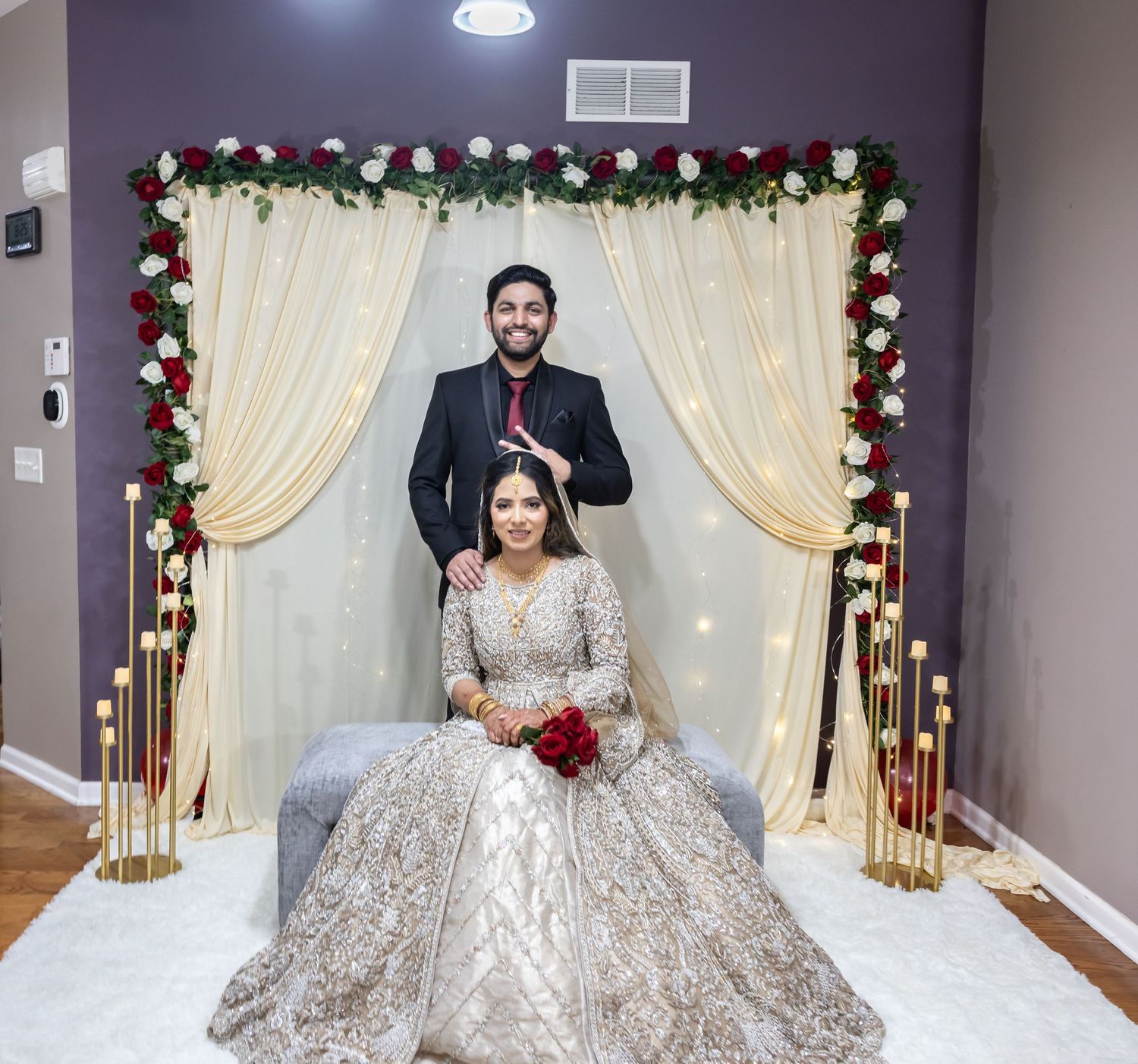 Bride and groom pose before cream drape arch adorned with red and white roses at indoor wedding.