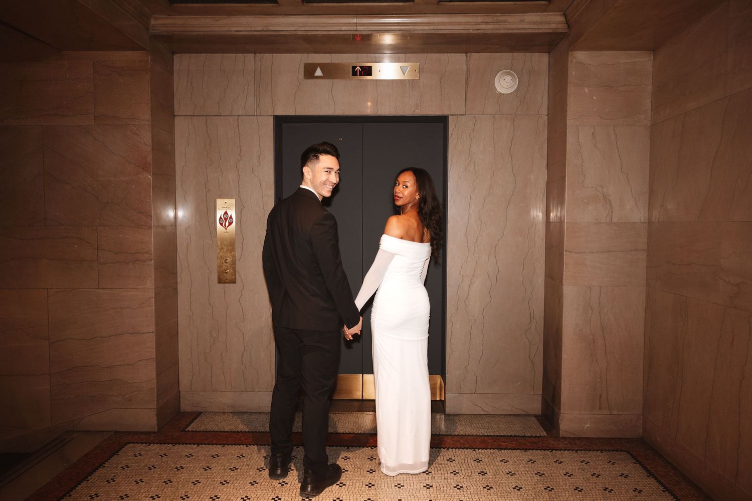 Couple holding hands tenderly outside a closed elevator, dressed in black suit and white bridal gown.Chicago Wedding Photographer