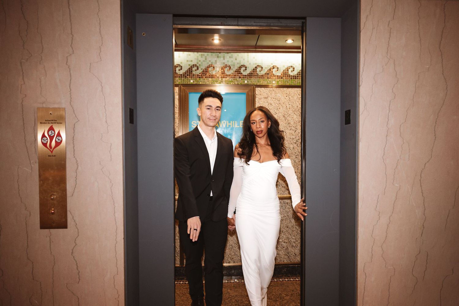 Smiling couple in black suit and white off-shoulder gown posing in decorative elevator doorway.Chicago Wedding Photographer