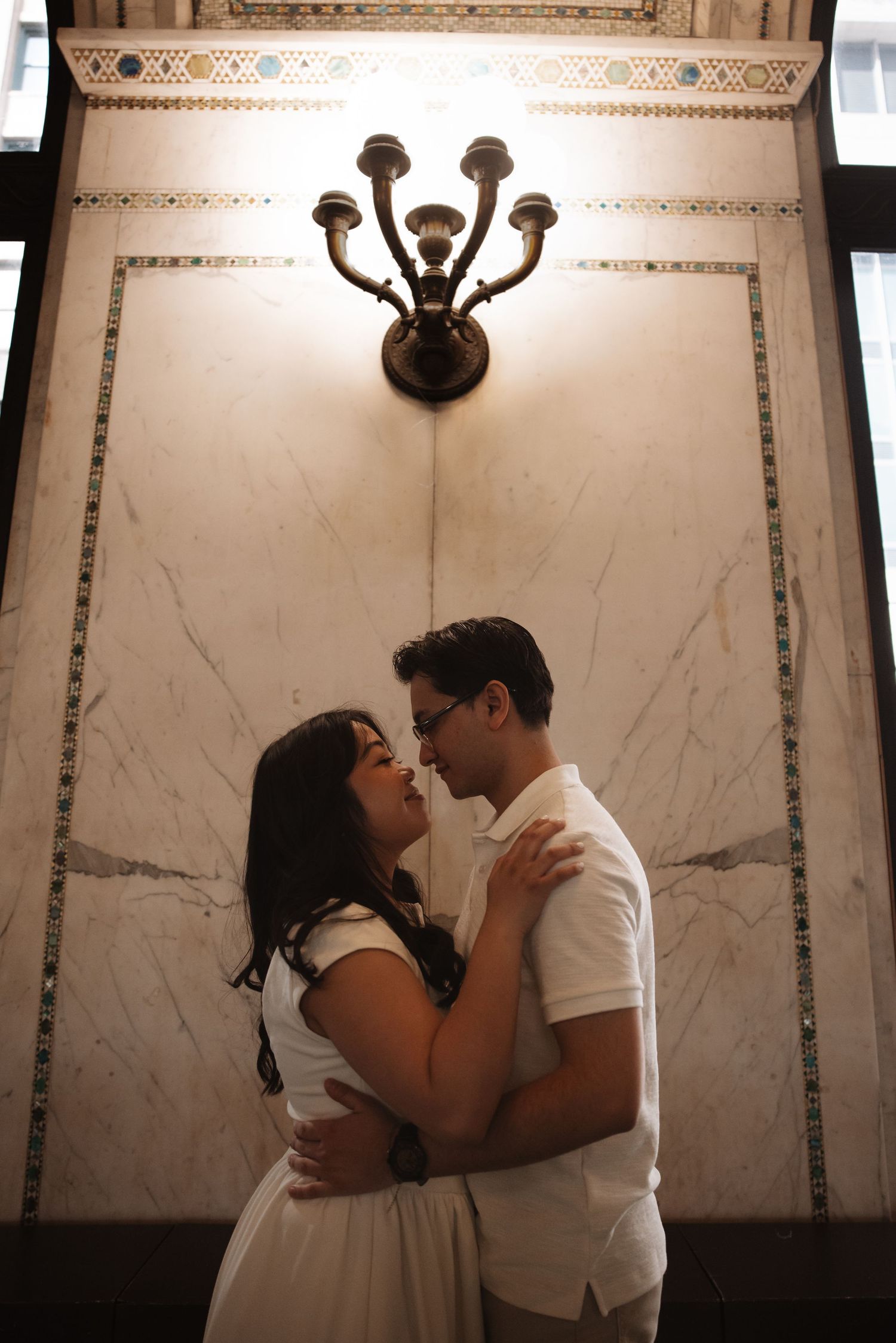 Couple shares tender moment against marble wall with vintage sconce.