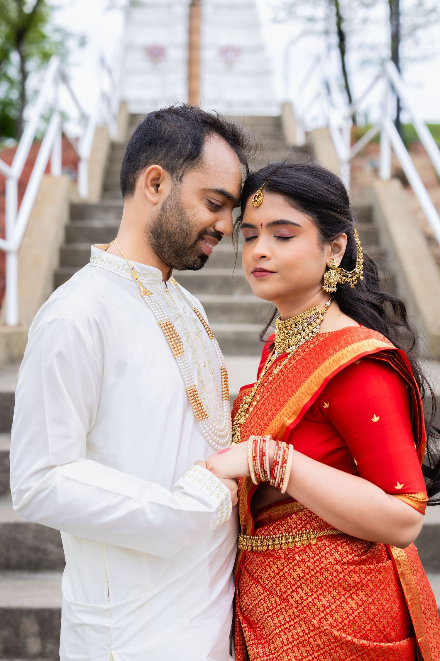 Indian couple in traditional wedding attire, bride in red saree with gold jewelry, groom in white kurta, holding hands.