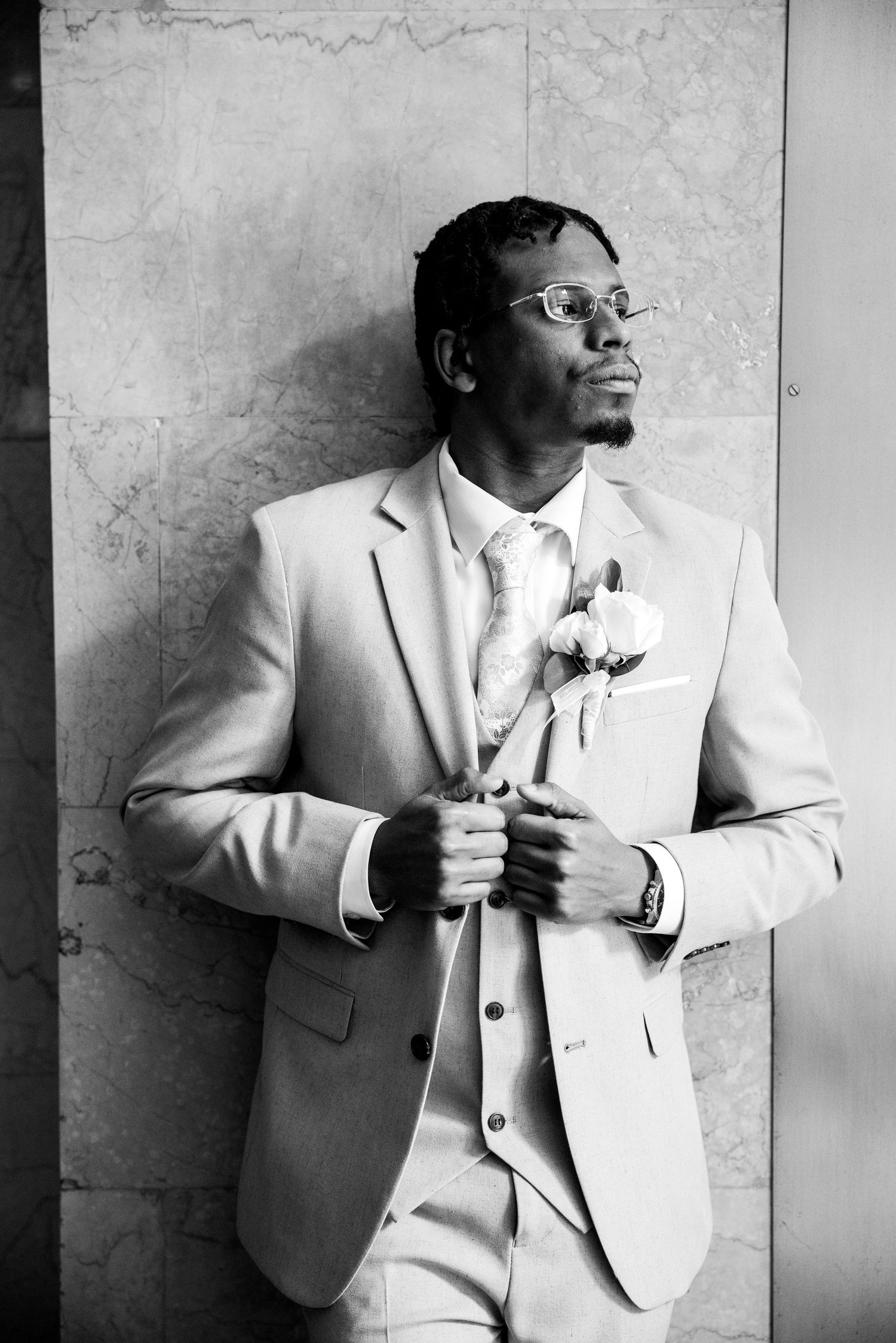 Elegant groom in light suit holding boutonniere flowers, leaning against marble wall in black and white portrait.