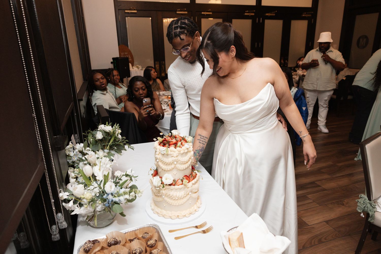 Bride and groom cut white wedding cake with strawberries surrounded by guests at reception celebration.