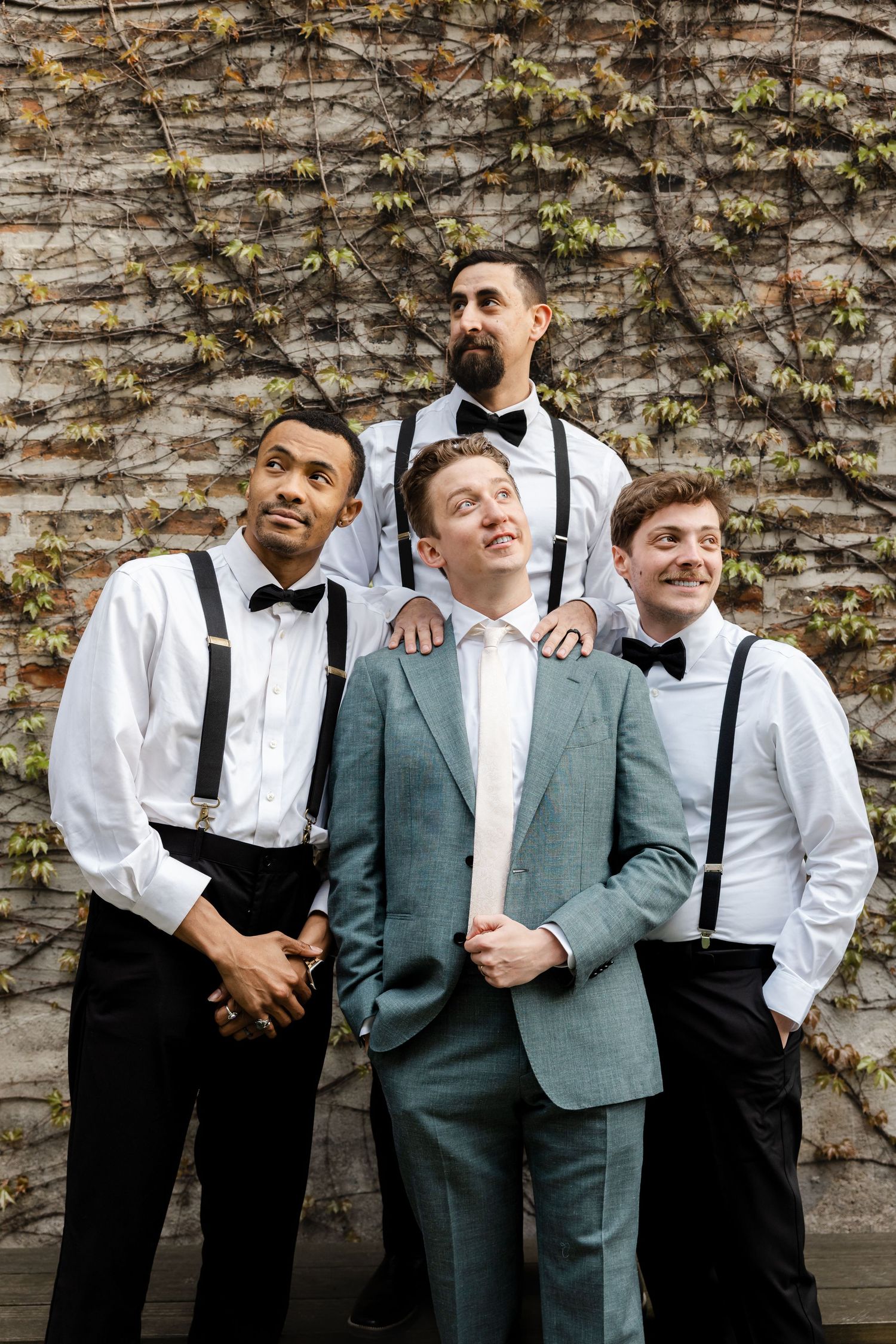 Groom in teal suit poses with three groomsmen in white shirts, bow ties, and suspenders against ivy-covered brick wall.