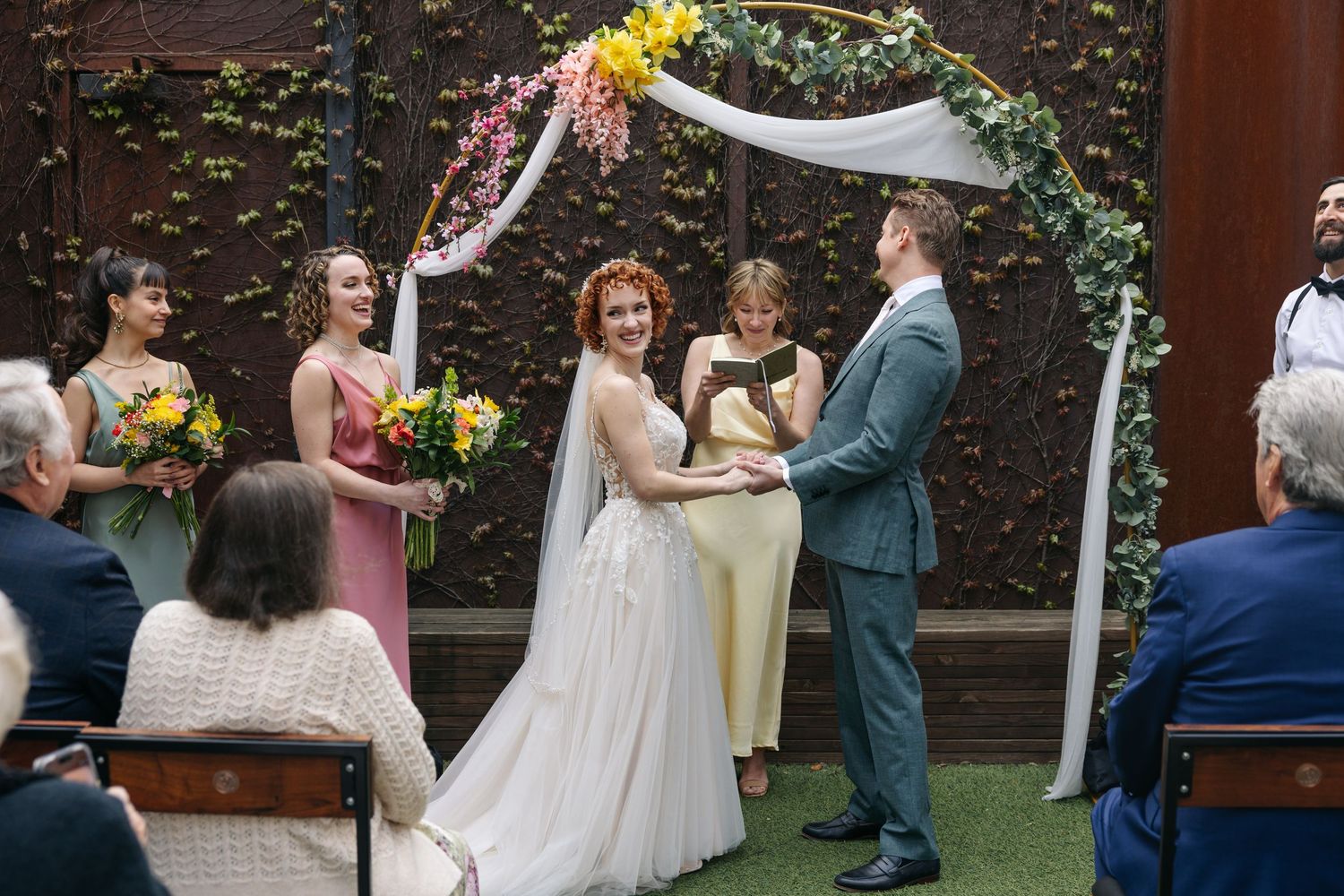Bride and groom exchange vows under floral arch with bridesmaids and guests at indoor wedding ceremony.