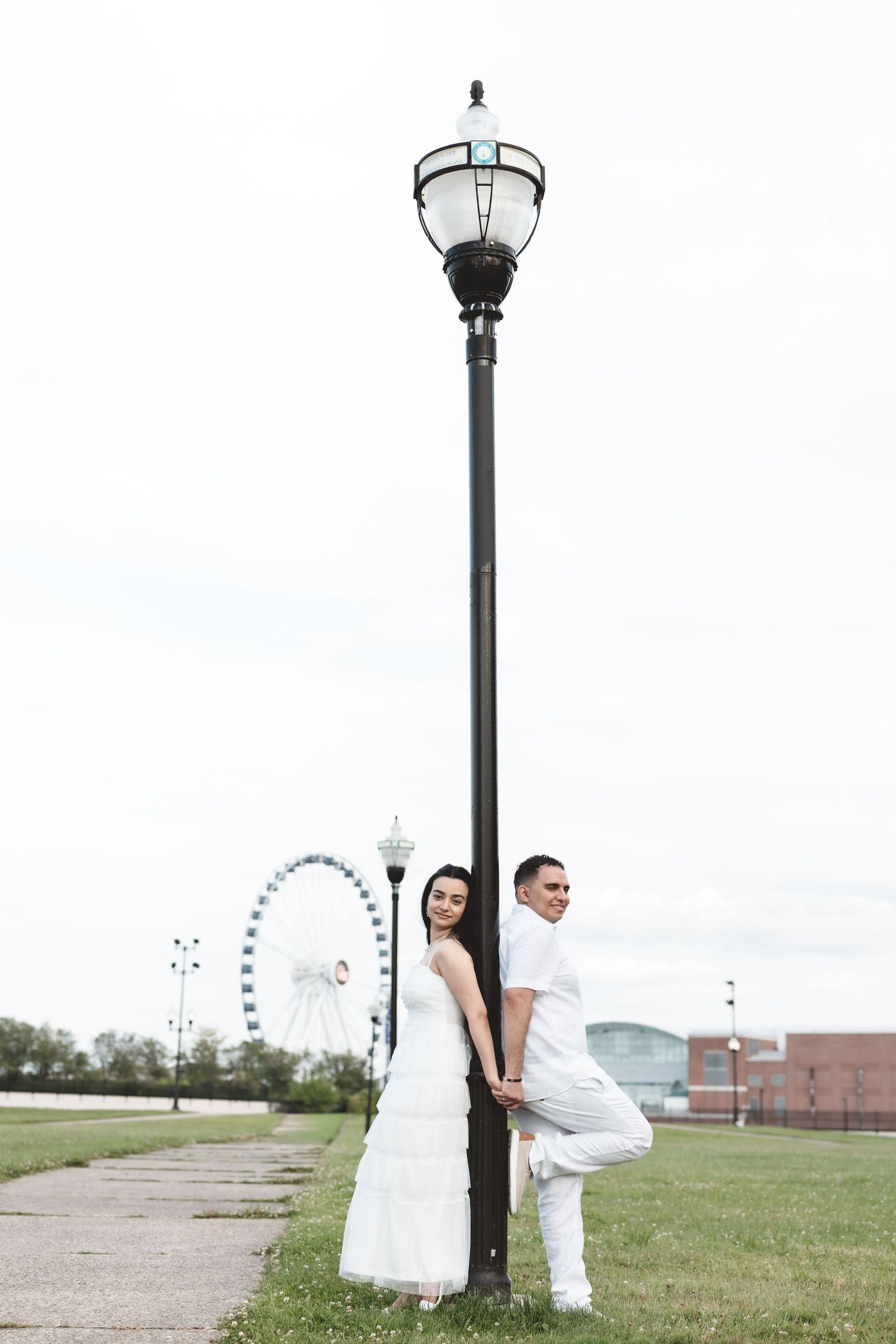Bride and groom in white posing by a lamp post with a Ferris wheel in the background.