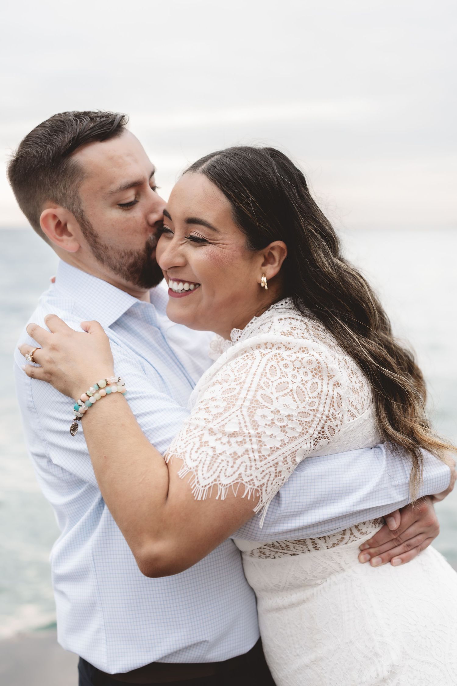 Groom kissing smiling bride on cheek while embracing near waterfront, woman in white lace dress with bracelet.