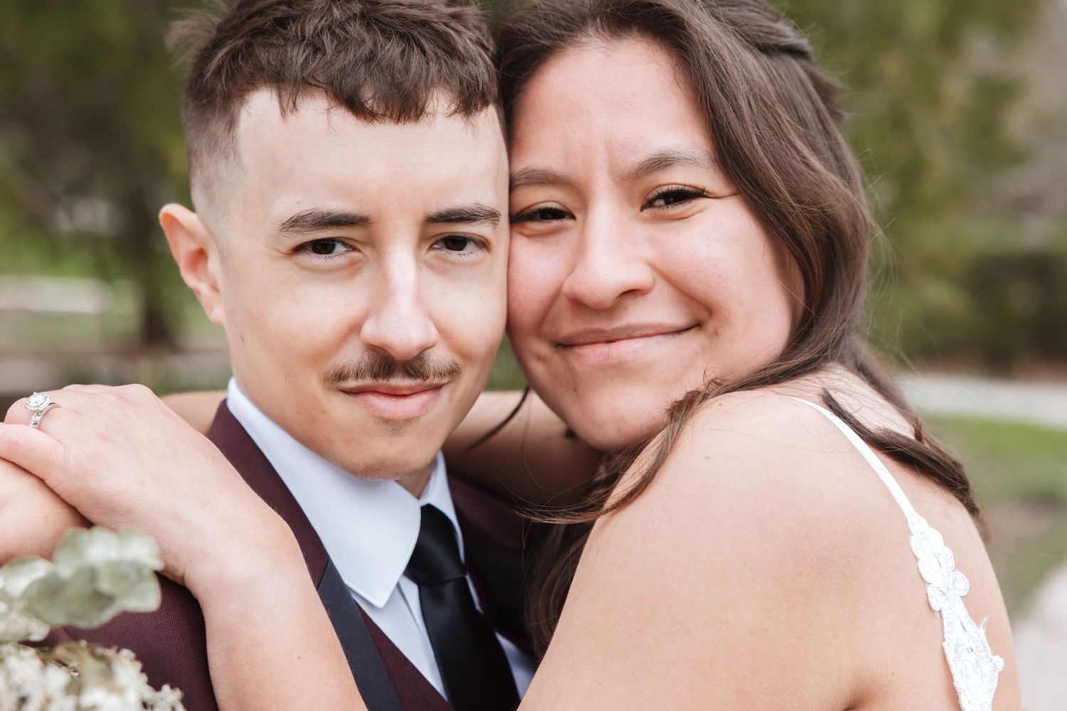A couple in wedding attire smiles closely together, the bride in white lace and groom in a dark vest and tie.