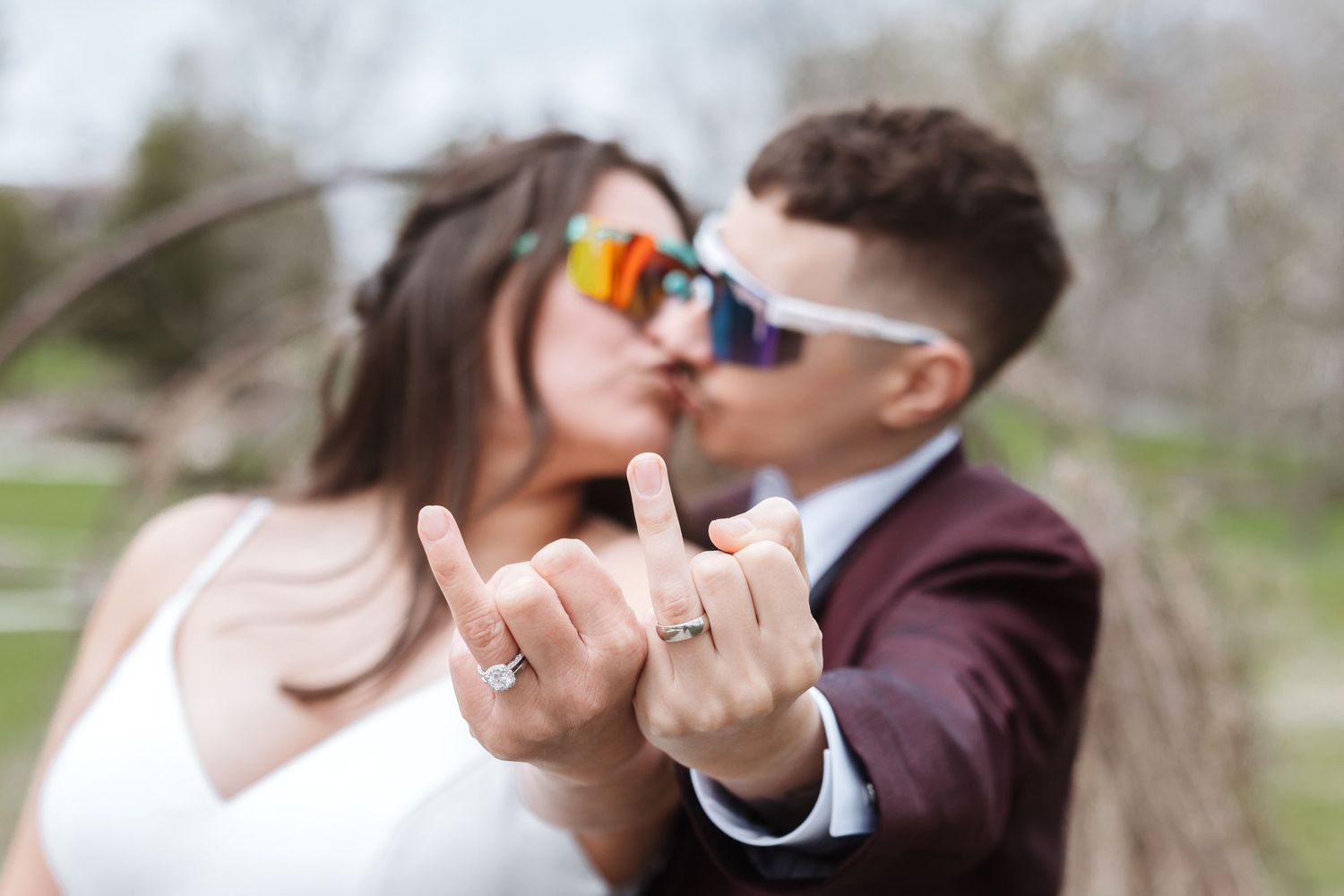 Newlywed couple kissing while showing off wedding rings, wearing stylish sunglasses outdoors on their wedding day.
