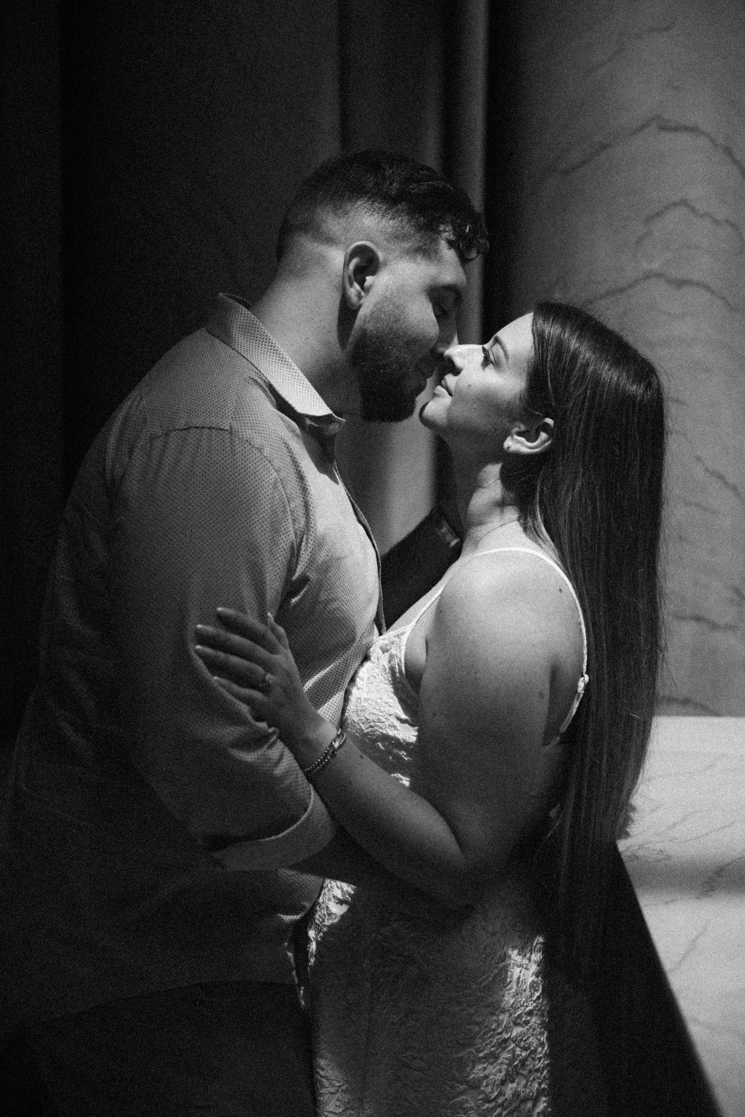 Black and white photo of couple sharing romantic almost-kiss embrace in dramatic moody architectural setting.