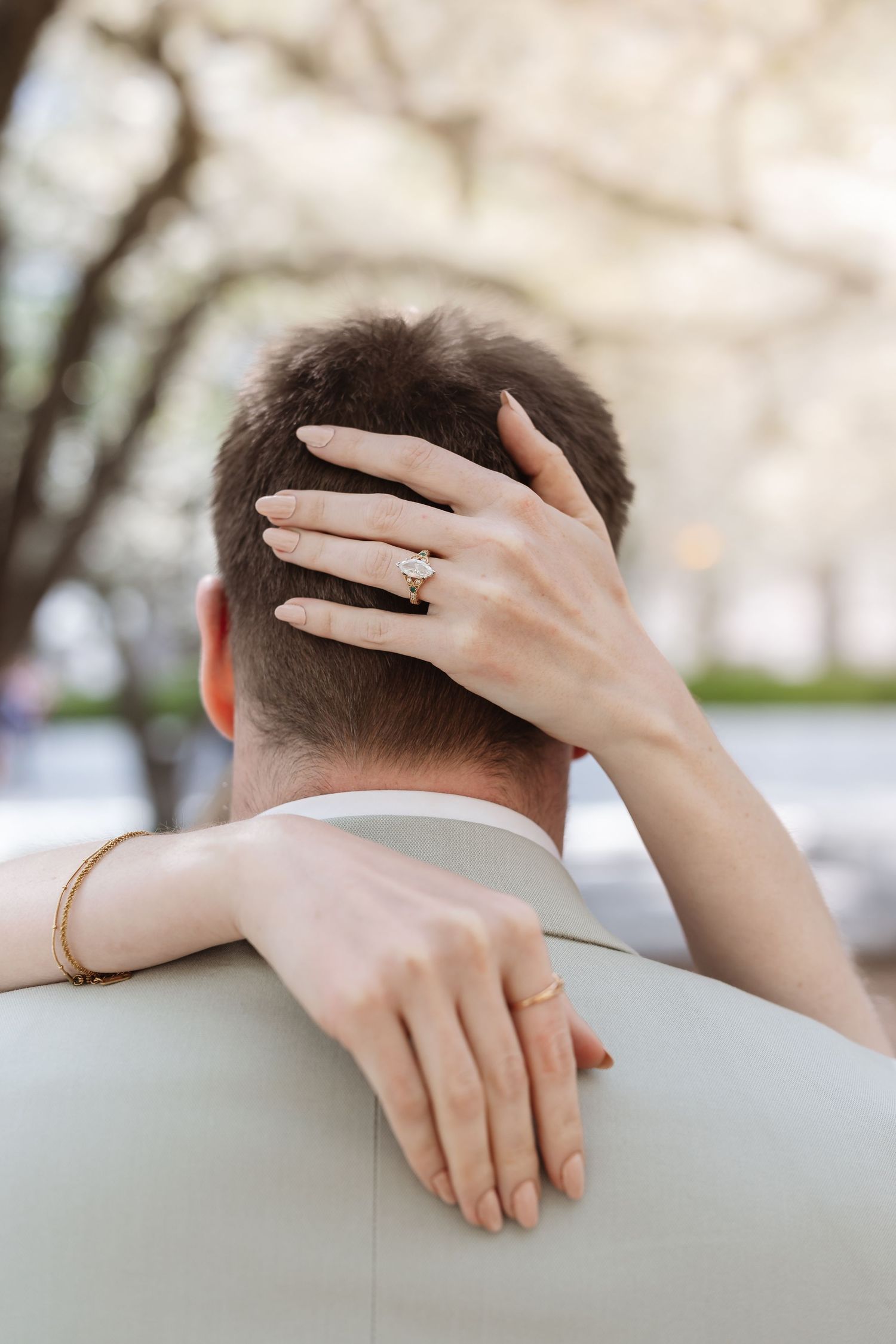 Close-up of bride's diamond engagement ring and gold bracelet as she embraces groom outdoors.