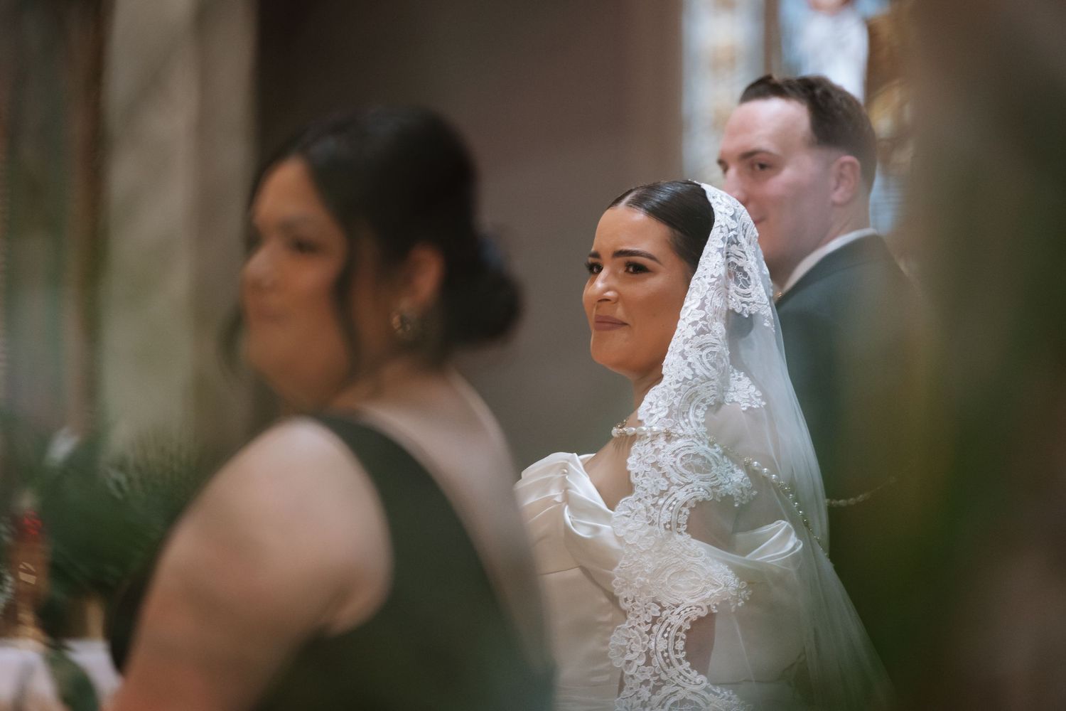 Bride wearing lace veil and gown stands with groom during wedding ceremony.