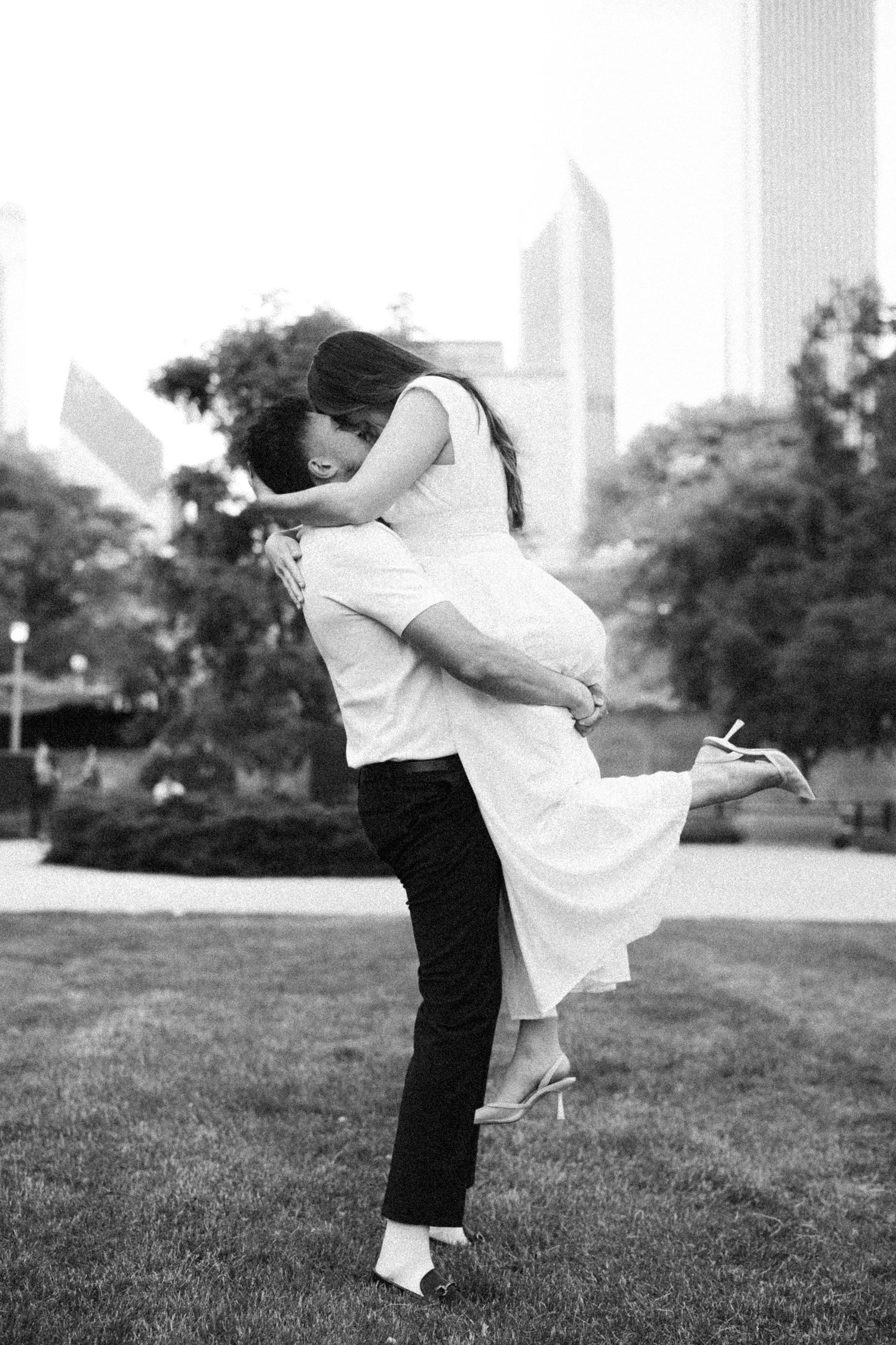 Black and white photo of a couple sharing a romantic kiss as the man lifts the woman in a park with city skyline.