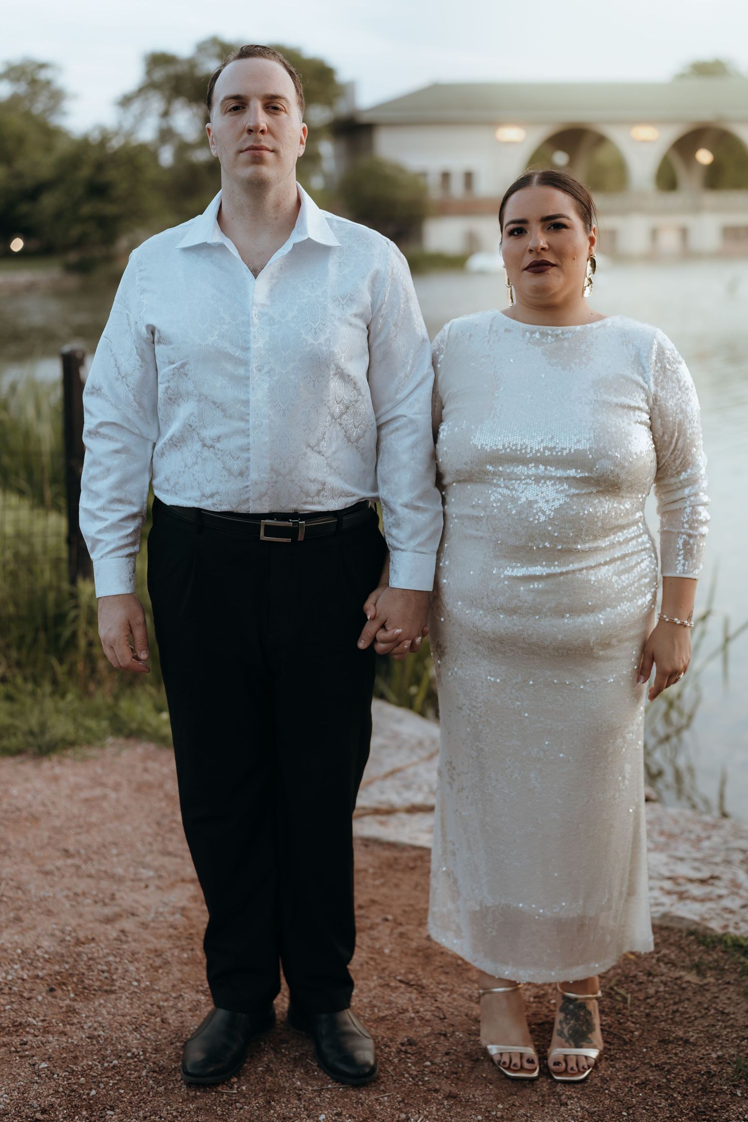 Couple posing hand in hand in coordinating white outfits beside a tranquil lake at dusk.