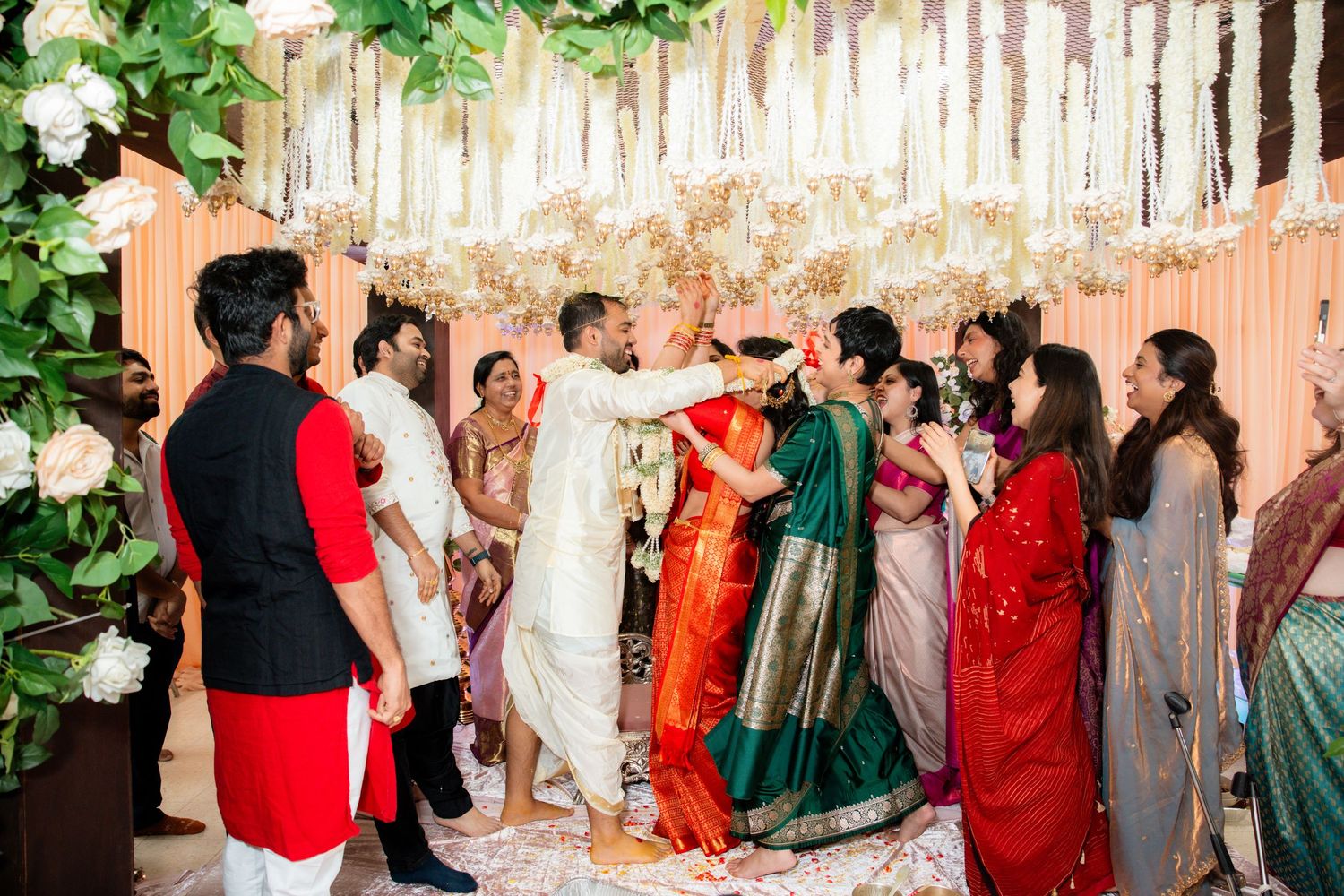 Indian wedding ceremony with groom in white garland exchange surrounded by guests in colorful traditional attire.