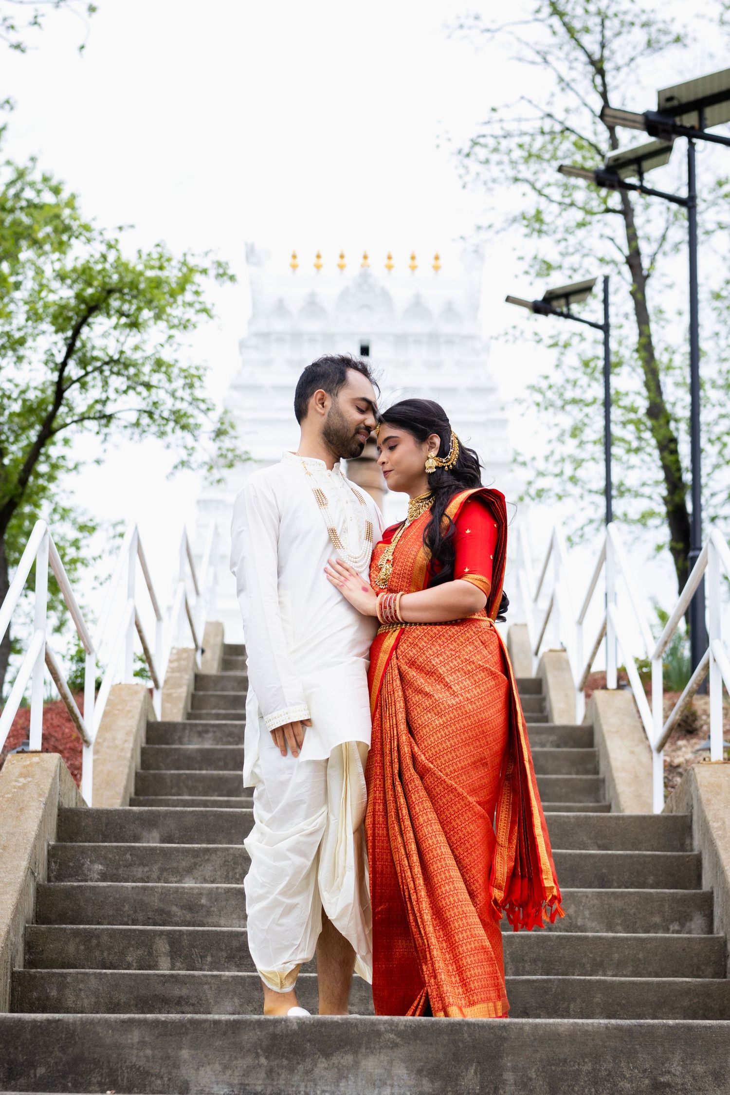 Indian bride and groom sharing an intimate moment on temple stairs in traditional red saree and white kurta.