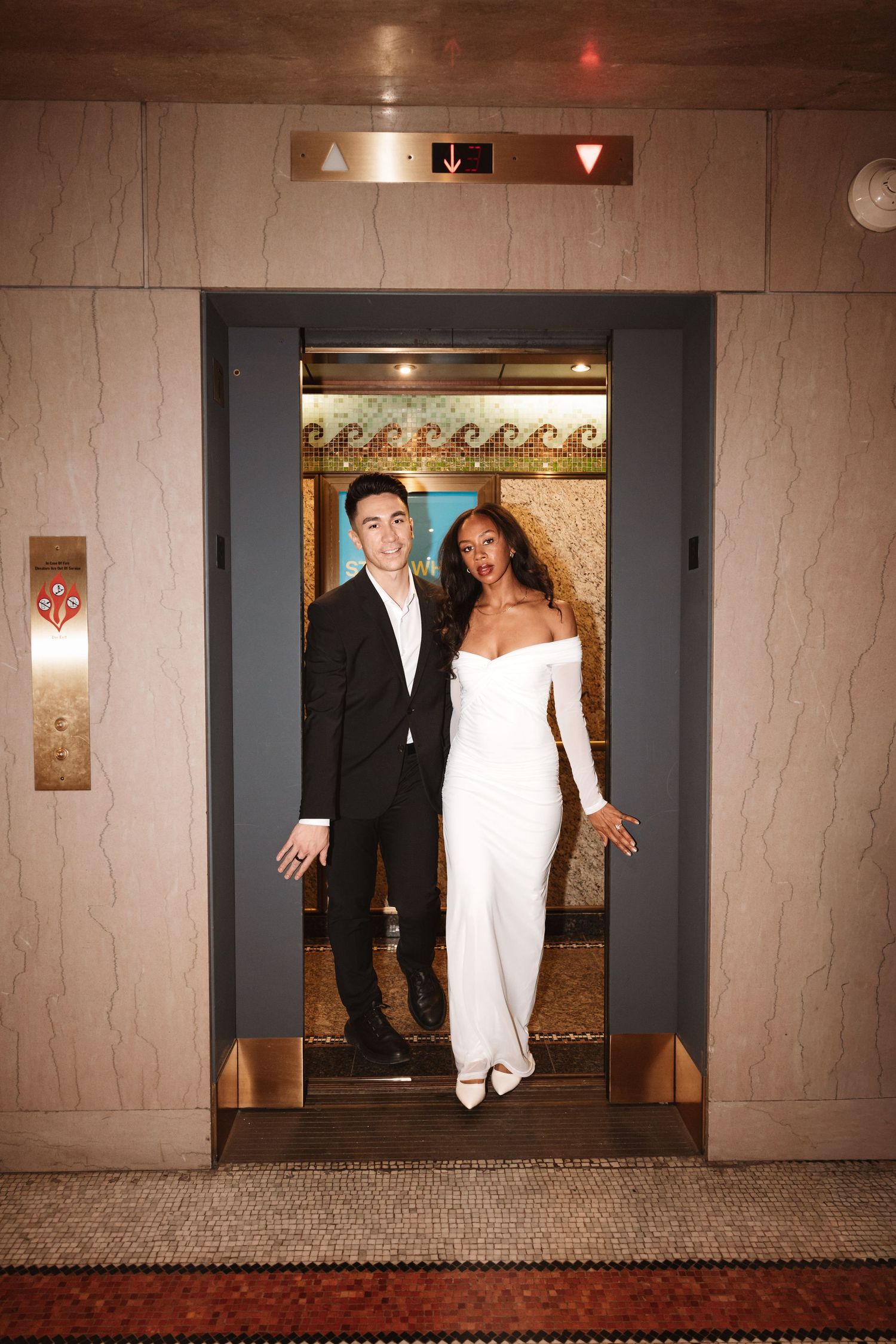 Joyful couple with arms outstretched standing in ornate elevator, wearing black suit and white gown.