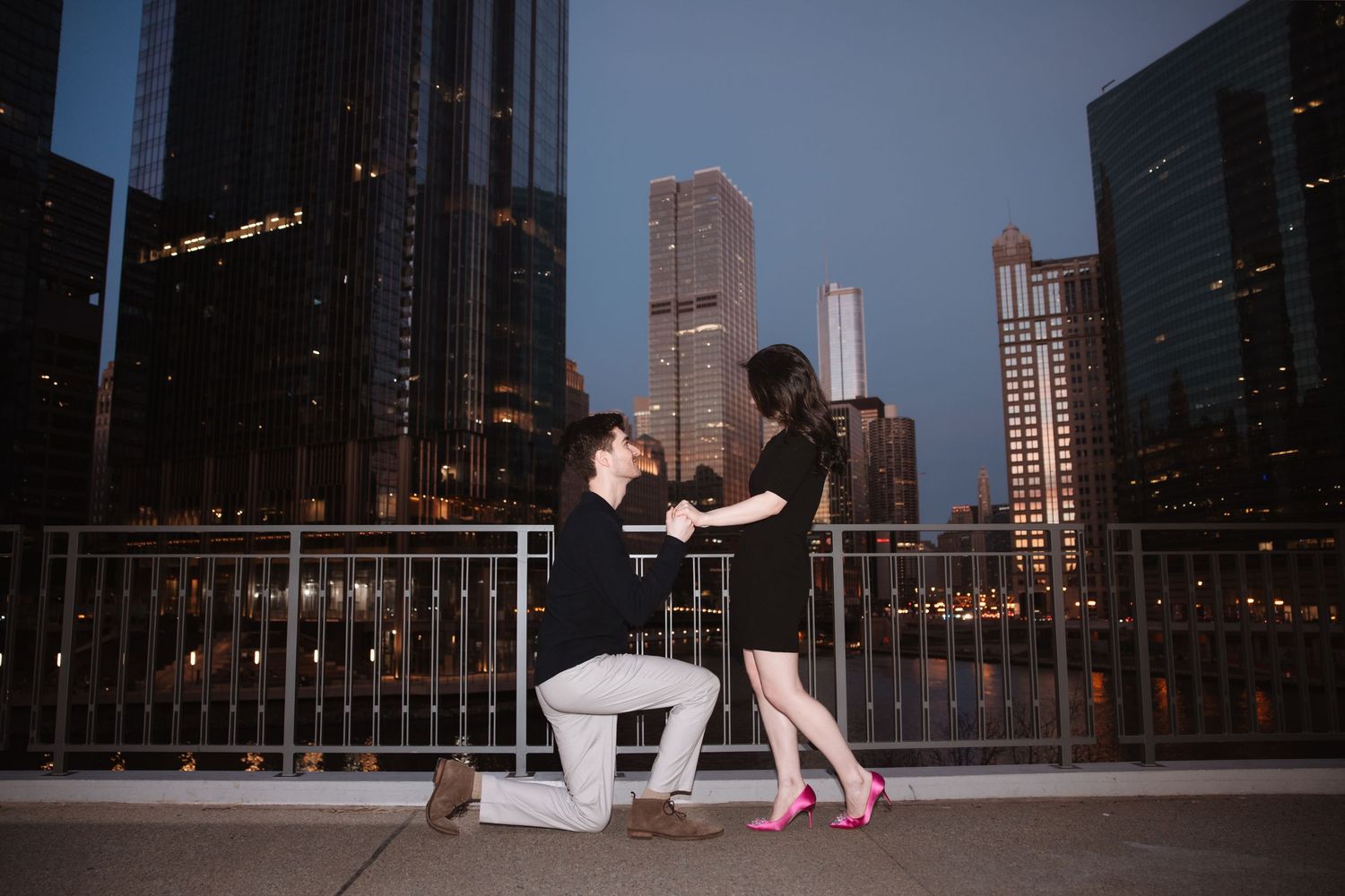 Man proposes on one knee to his girlfriend on a Chicago bridge at dusk with city skyline in the background.