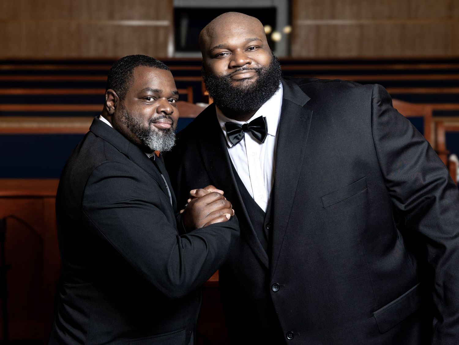 Two men in black tuxedos sharing a handshake and smiling inside a church setting.
