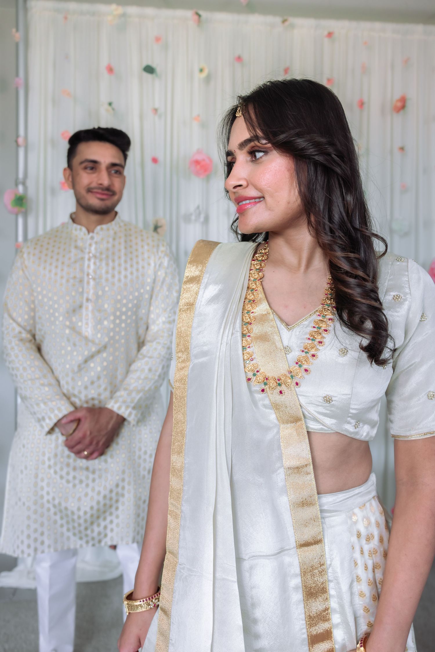 South Asian bride in white lehenga with gold trim smiling as groom stands in background.
