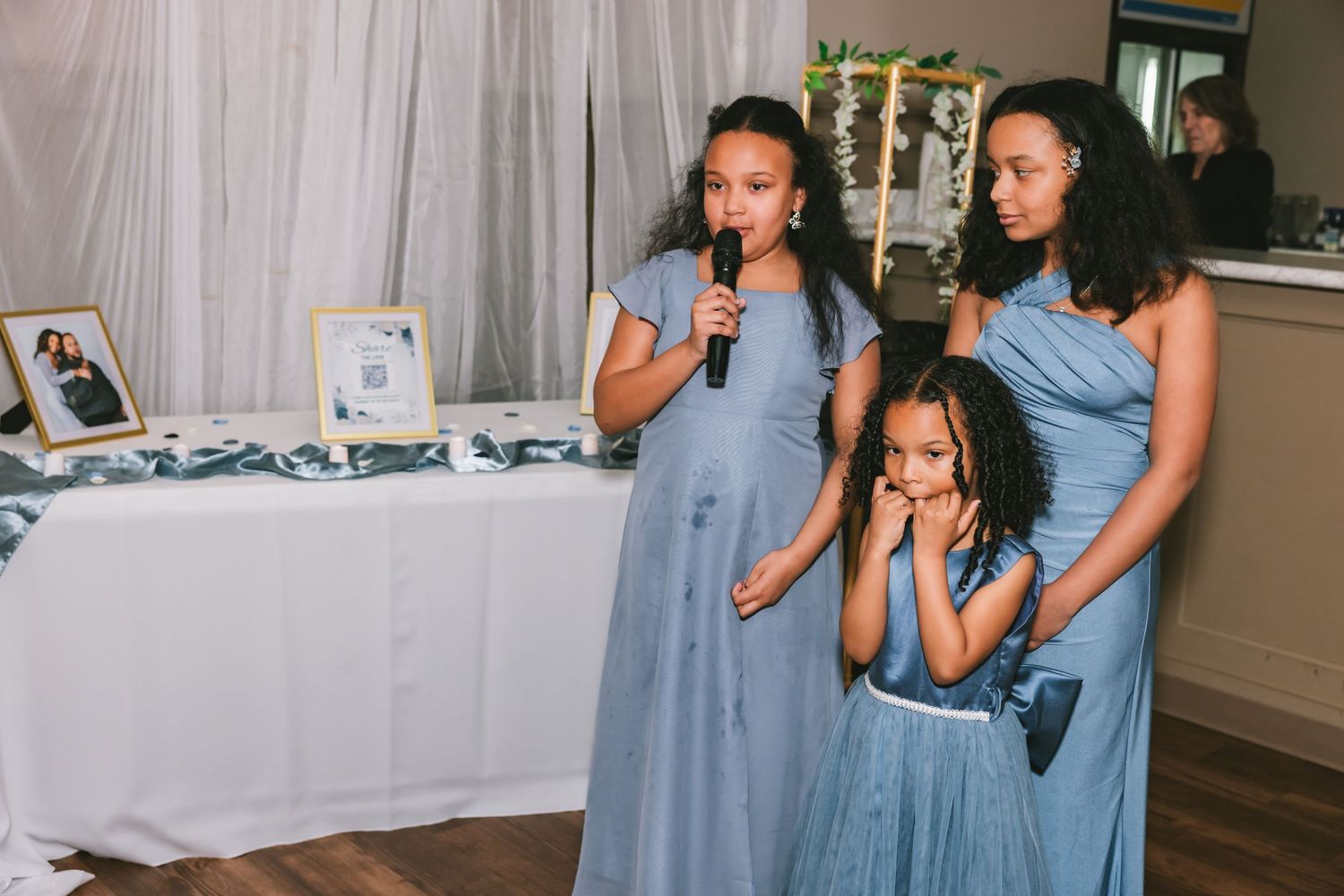 Three girls in blue dresses, one speaking into a microphone at a wedding reception.