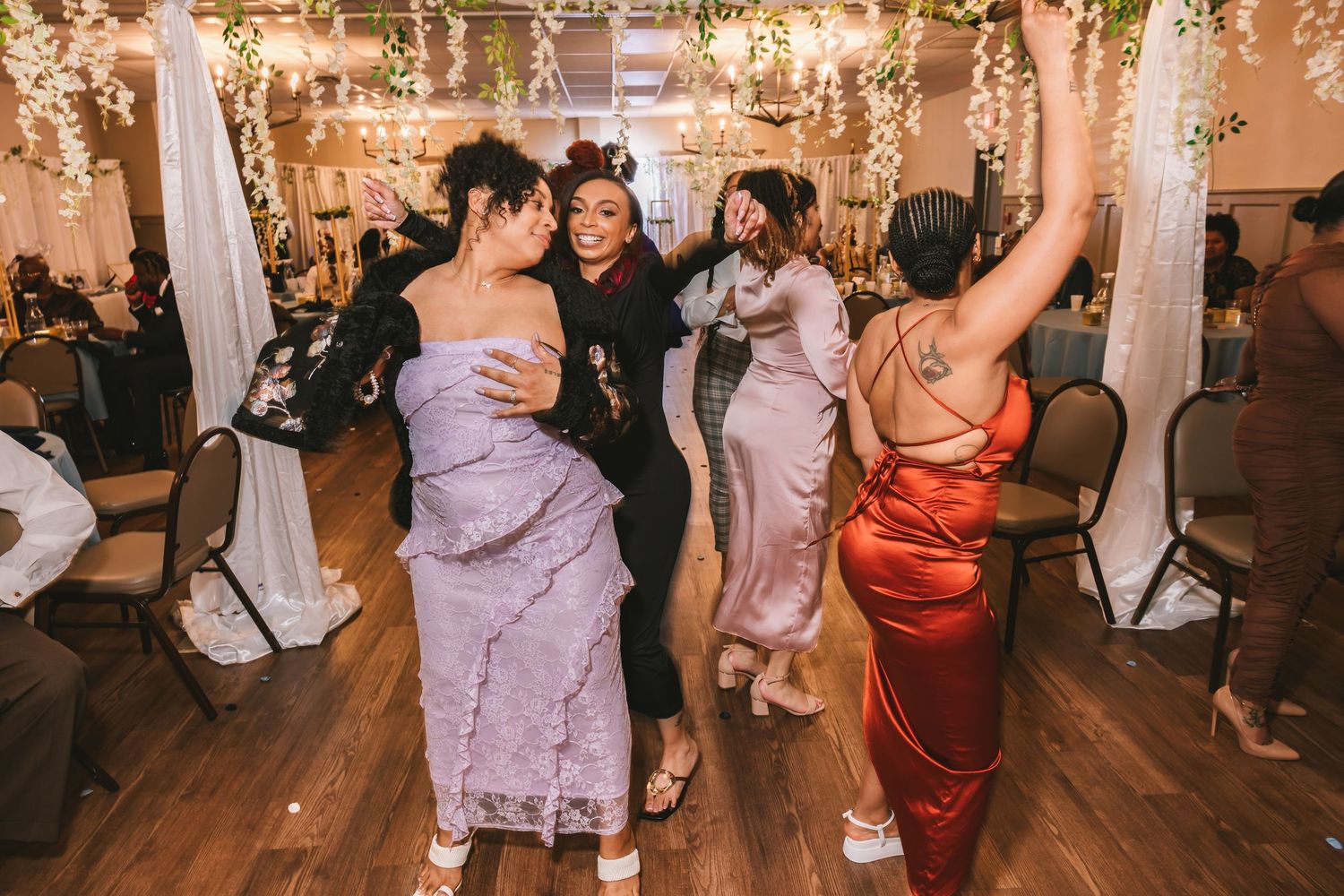 Four women in elegant dresses dancing joyfully at a wedding reception with string lights overhead.