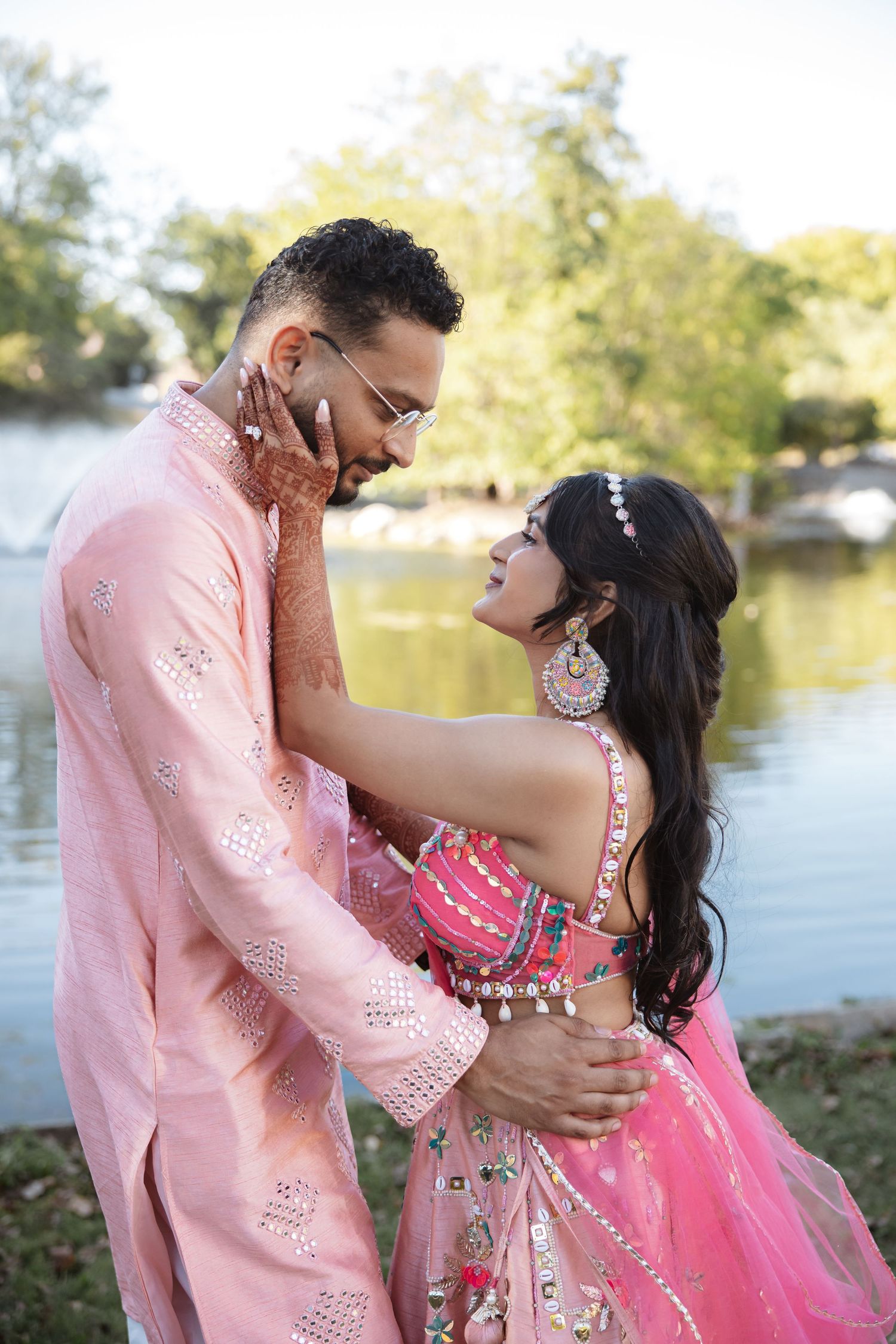 Indian couple in pink traditional attire embrace romantically by a lake, bride adorned with mehndi and silver jewelry.