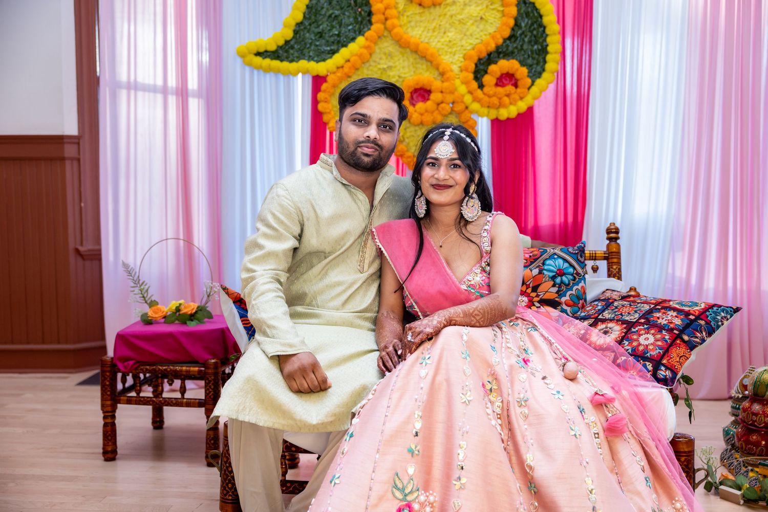 Indian couple in pink traditional wedding outfits pose at colorful mehendi ceremony.