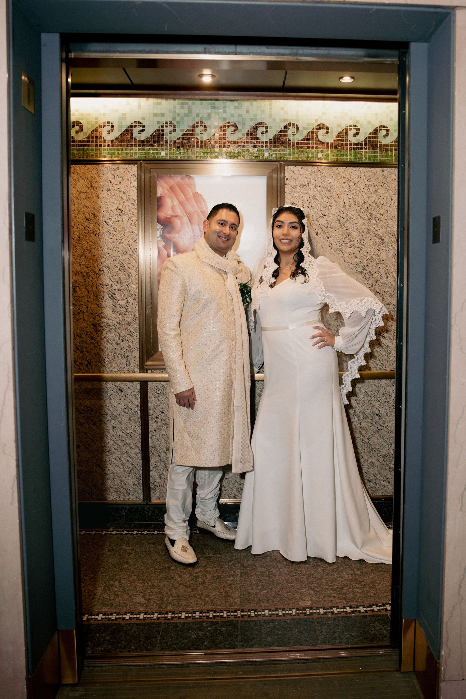 Bride and groom pose smiling inside an ornate elevator with mosaic tile details.