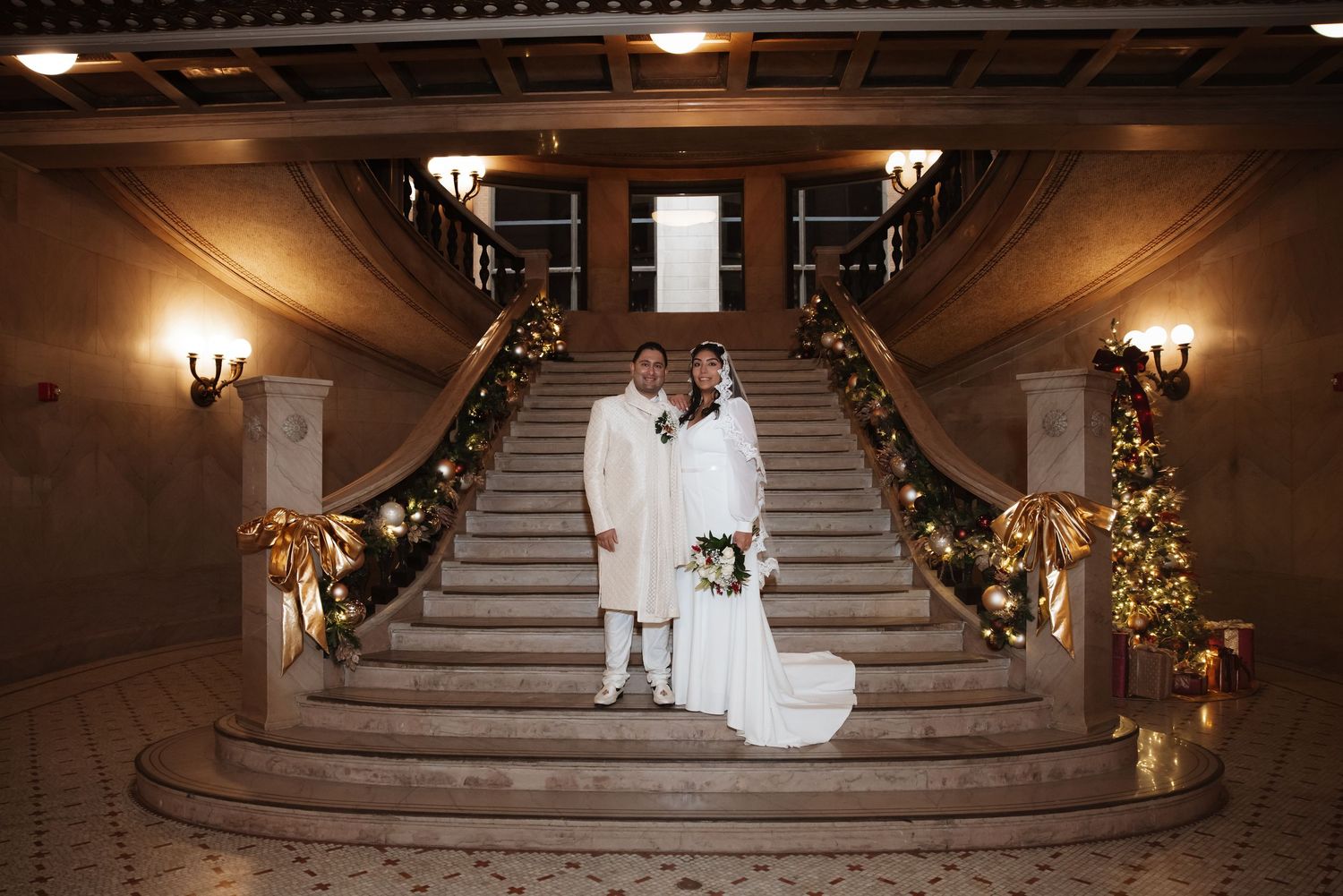 Wedding couple posing on sweeping marble staircase decorated with holiday greenery and warm ambient lighting.