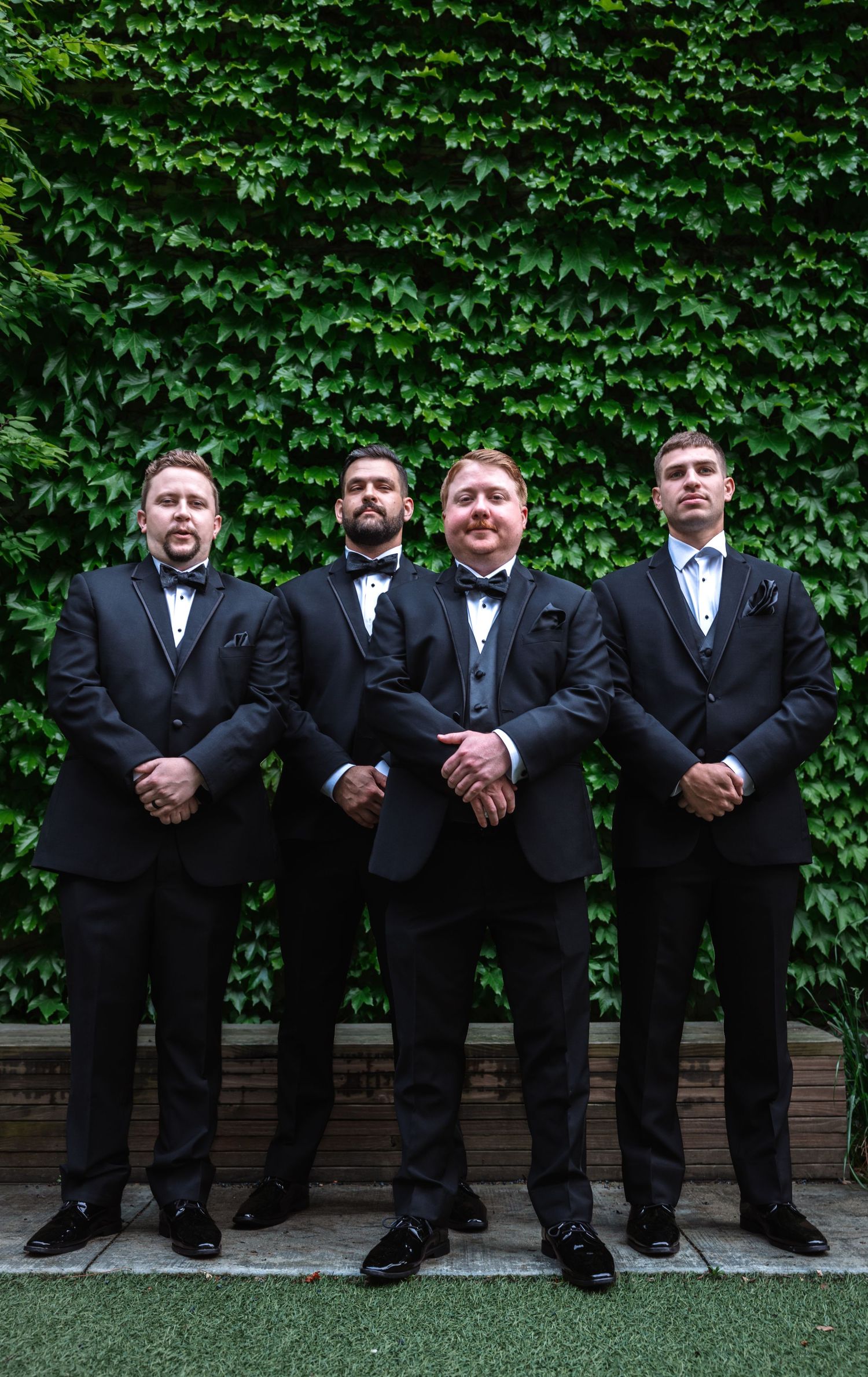 Four groomsmen in black tuxedos standing formally in front of a lush green ivy wall.