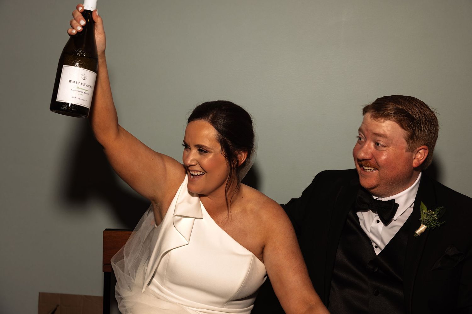 Excited bride in white dress raises champagne bottle while groom in tuxedo laughs beside her at wedding reception.