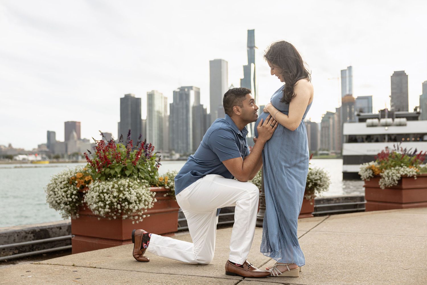 Man on one knee kissing pregnant partner's belly near Chicago lakefront with urban skyline backdrop.
