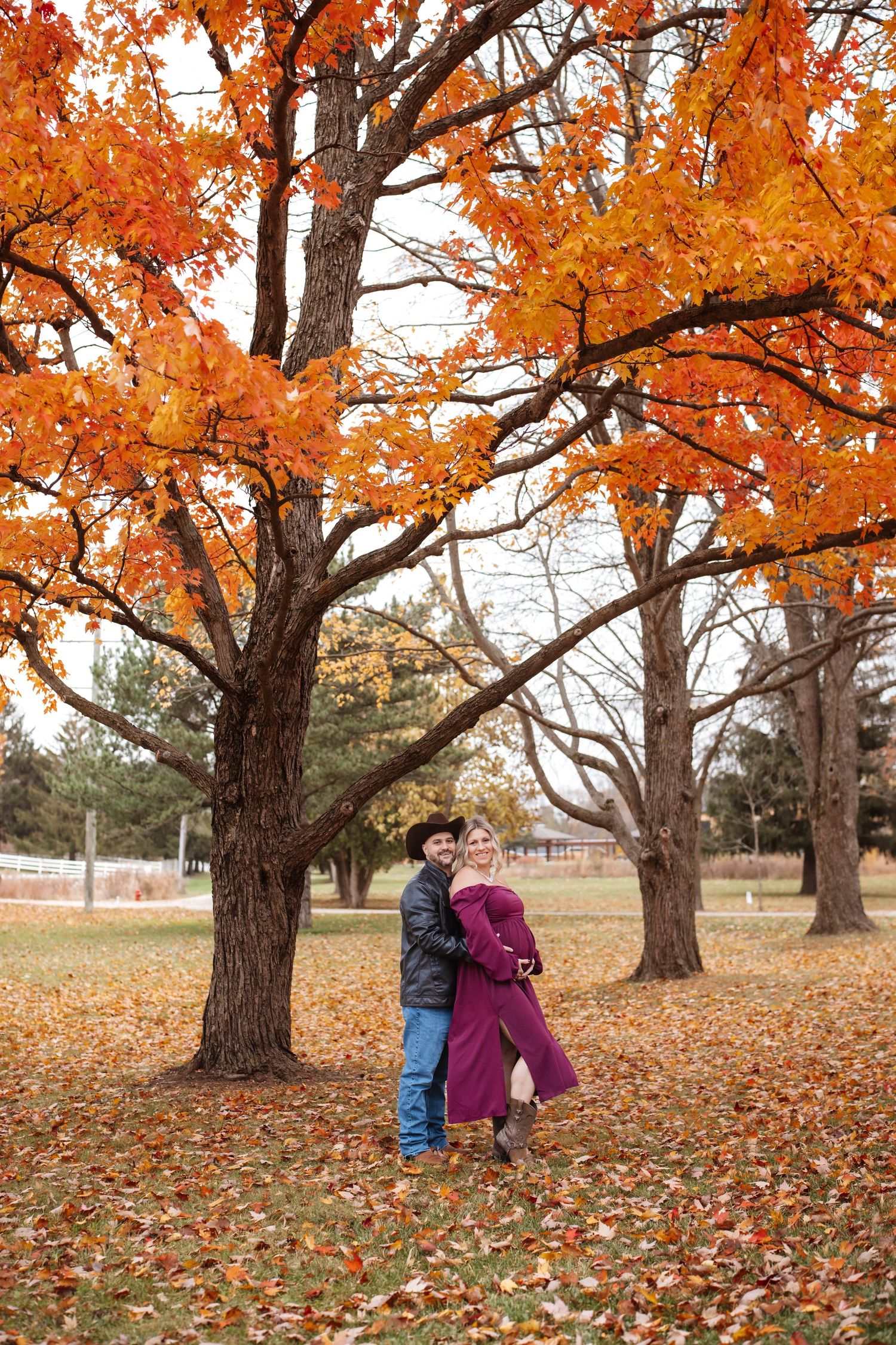 Couple embracing under vibrant orange autumn trees, man in cowboy hat with pregnant woman in burgundy maternity dress.
