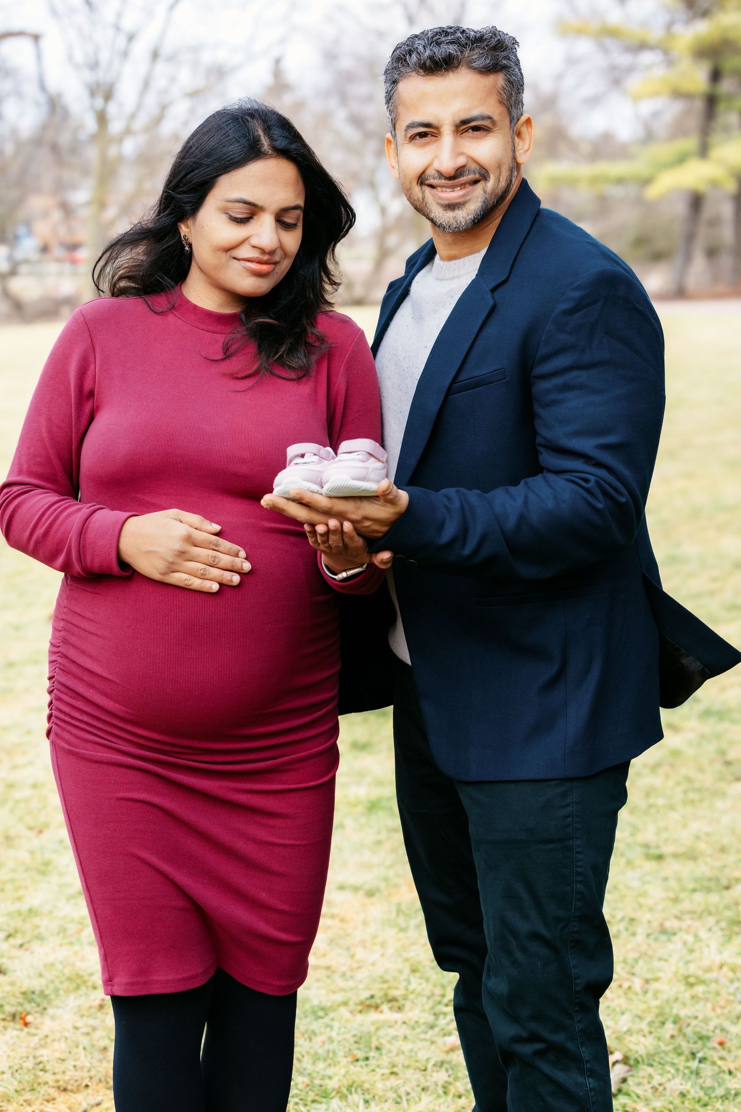 Smiling expecting couple in a park holding small pink baby shoes, woman in red dress.