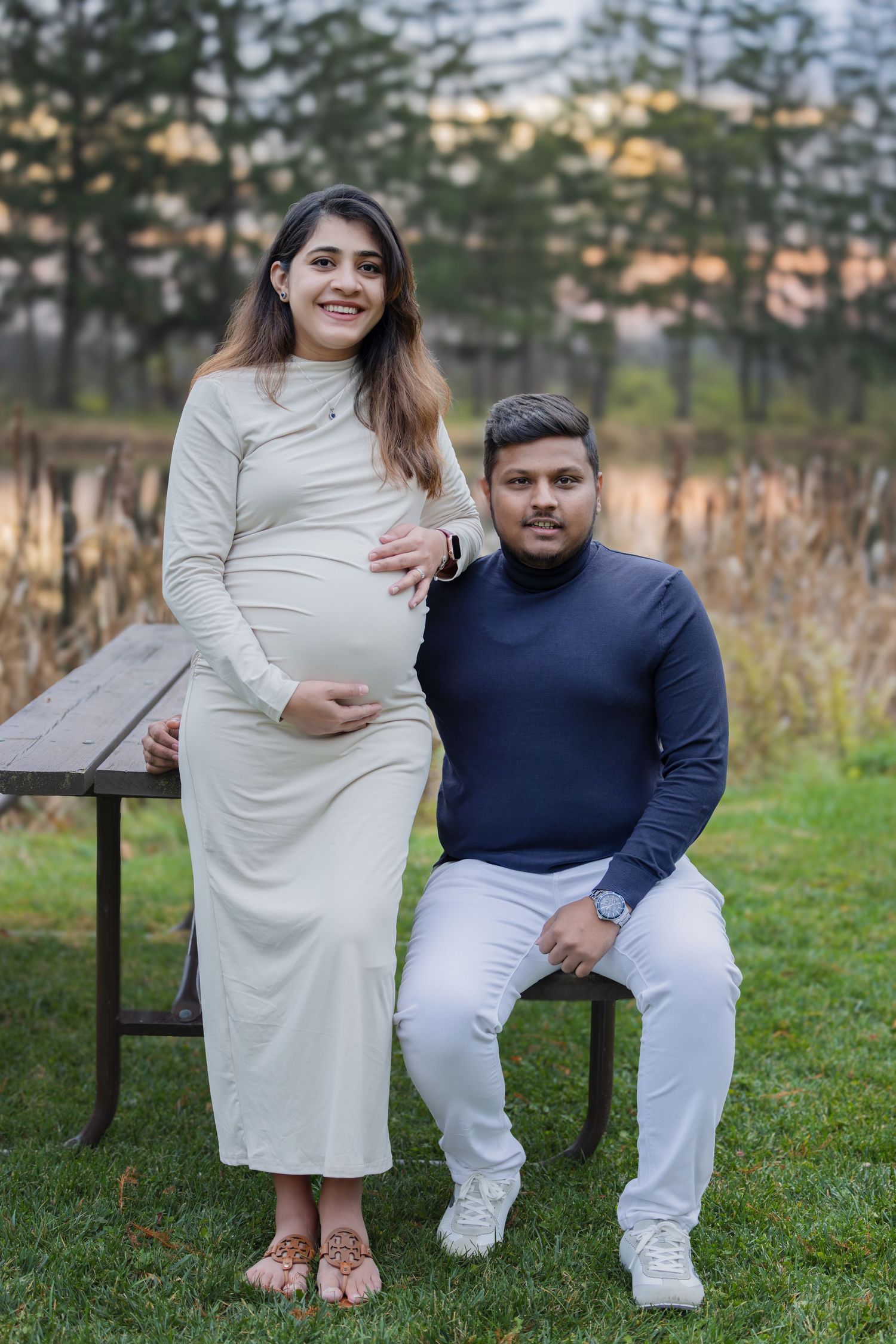 Smiling pregnant woman in white dress standing beside seated partner near pond at sunset.
