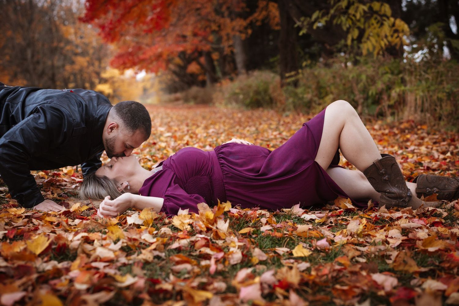 Couple sharing a tender kiss among vibrant fallen autumn leaves, pregnant woman in purple dress lying on the ground.
