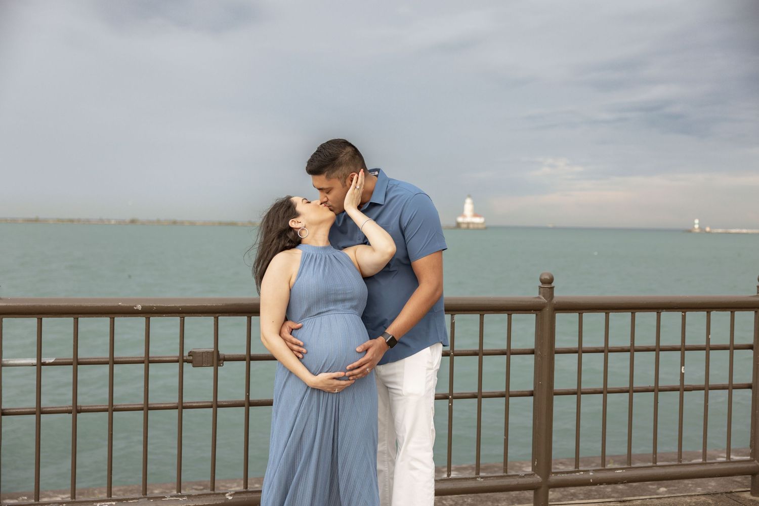 Pregnant couple sharing a romantic kiss on a lakeside pier with a lighthouse visible in the background.
