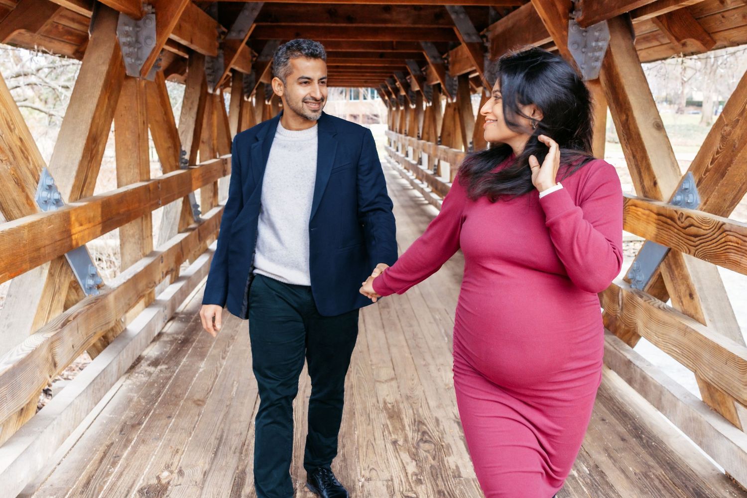 Pregnant couple walking hand in hand through a wooden covered bridge, woman in burgundy maternity dress.