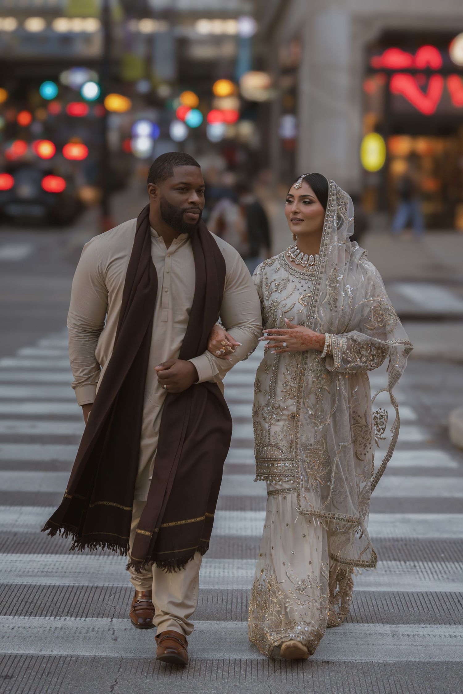 Interracial couple in traditional South Asian wedding attire walking hand in hand across a city crosswalk at dusk.