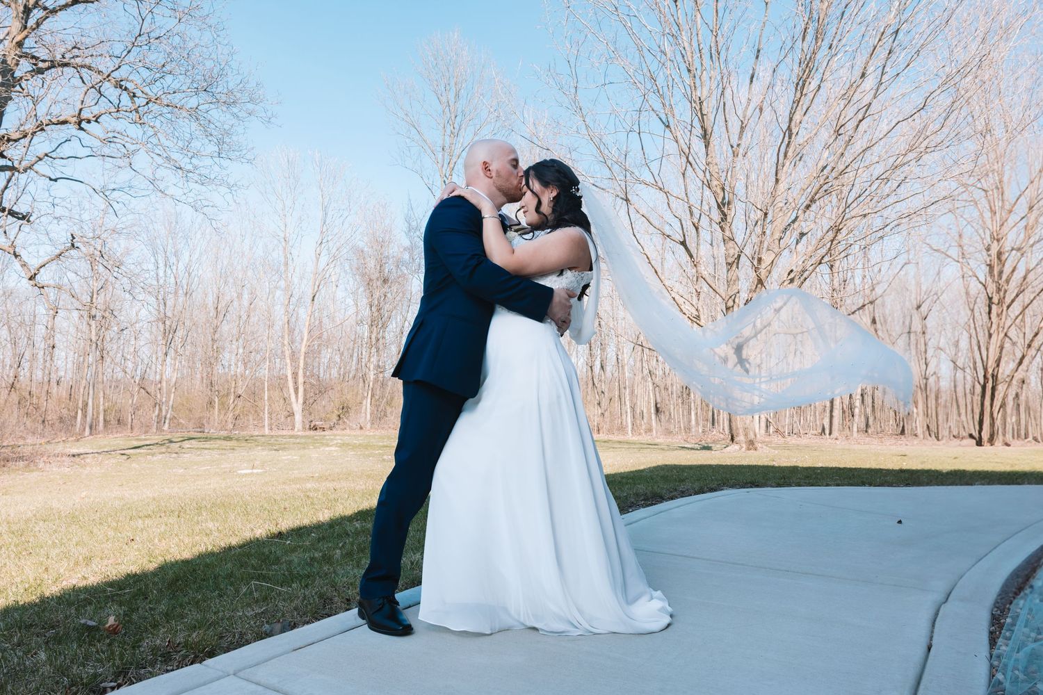 Bride and groom share a kiss outdoors with wedding veil flowing in the breeze.