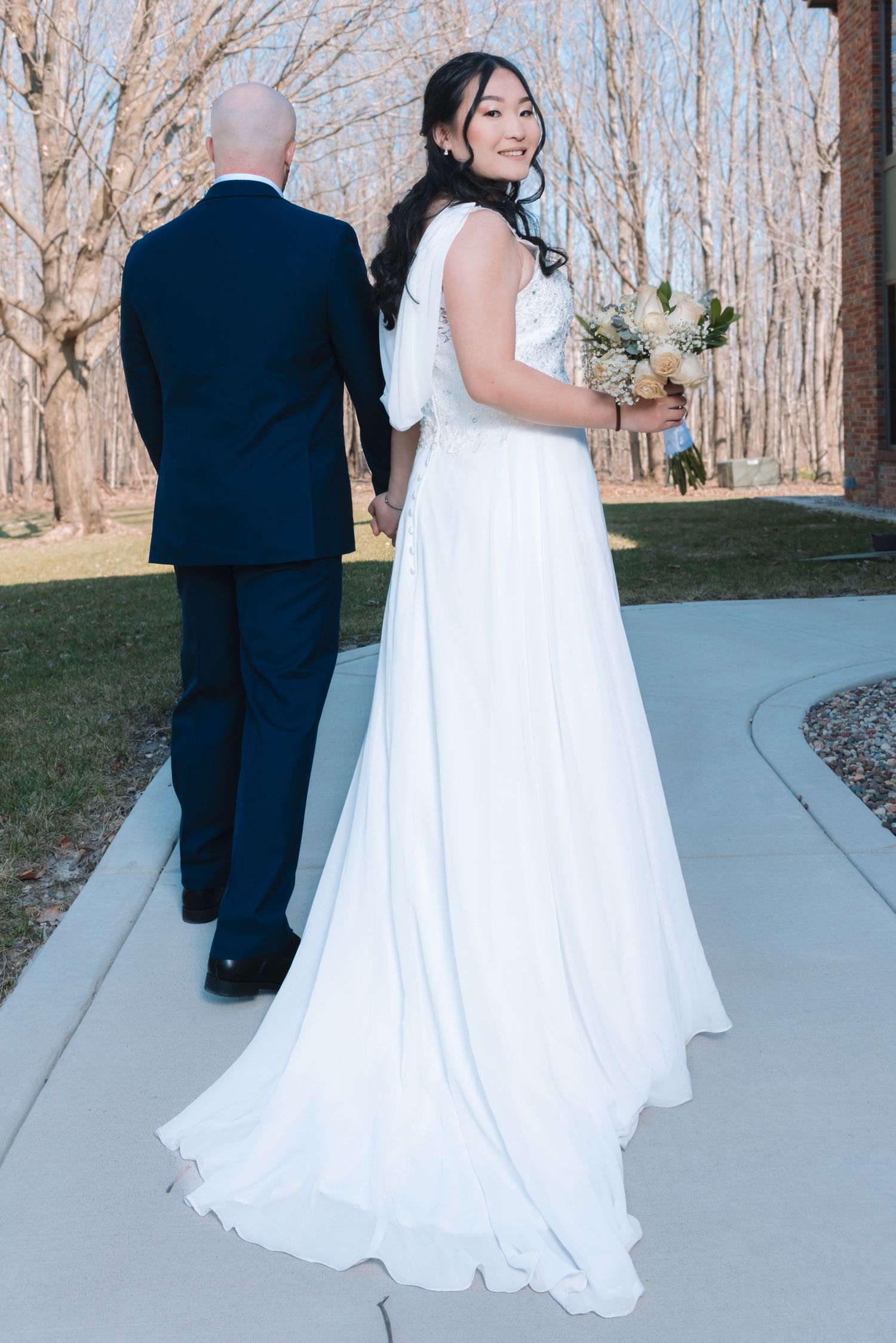 Smiling bride in elegant white gown holds bouquet while walking with groom on outdoor path.