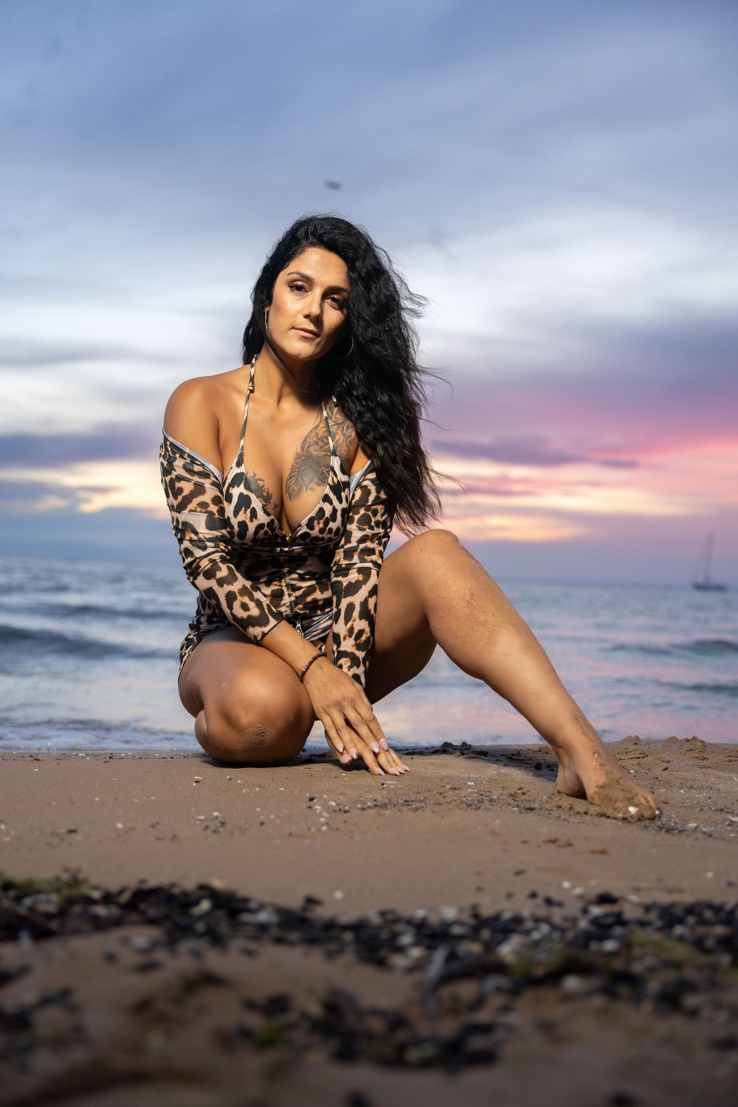 Curly-haired woman in leopard print swimwear sitting on rocky beach shore at dramatic sunset.