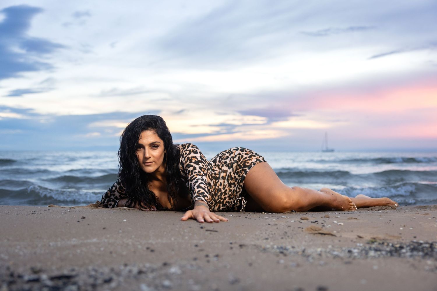 Woman in leopard print dress posing on beach sand at sunset with ocean and cloudy sky.