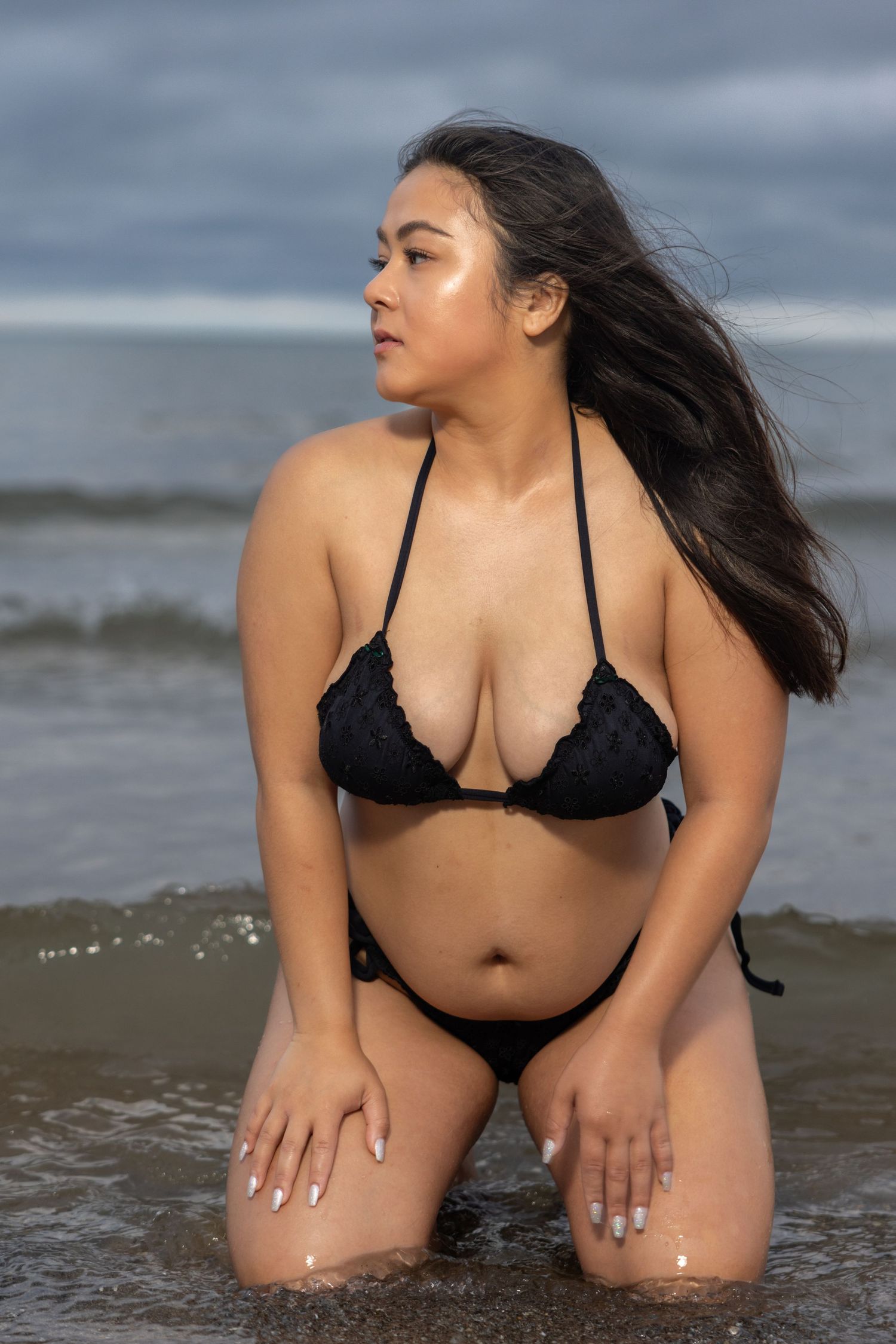 Woman in black lace bikini kneeling in shallow ocean waves with long dark hair blowing in wind.