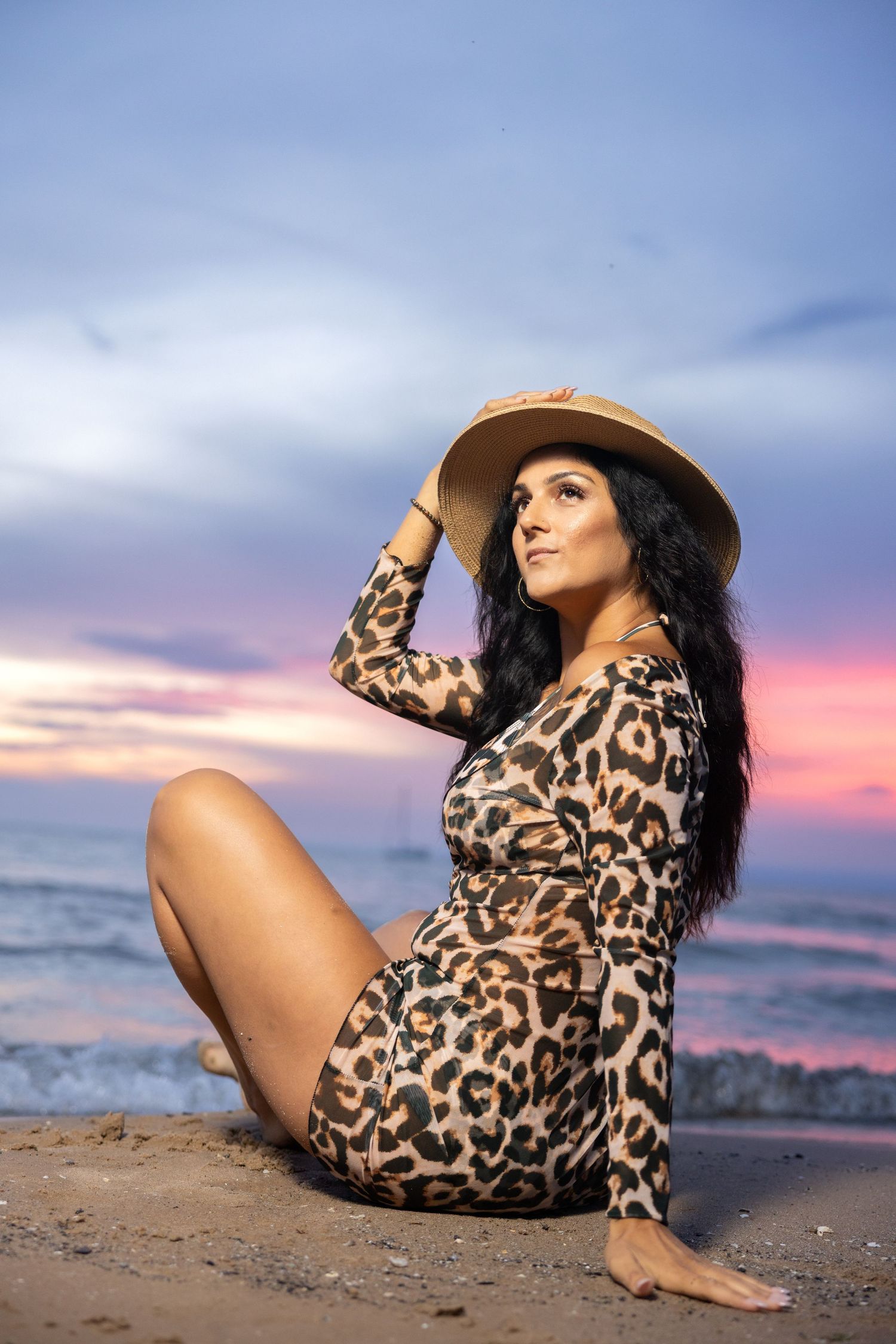 Brunette woman in leopard outfit holding straw hat on beach at dusk.