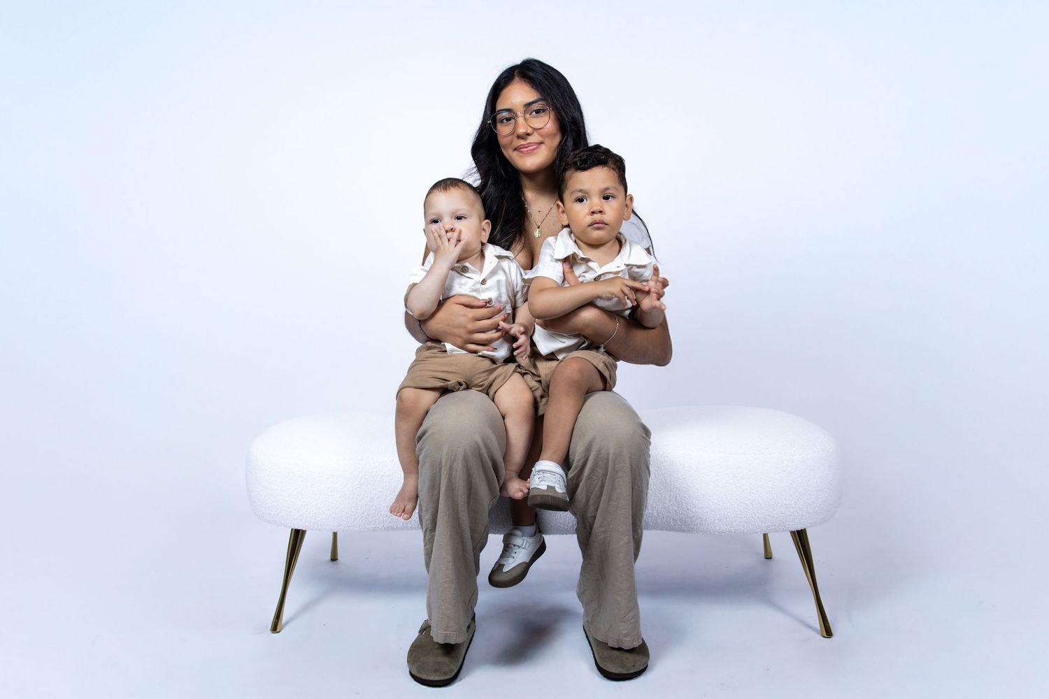 Young mother sits on white bench holding twin babies in matching beige outfits.